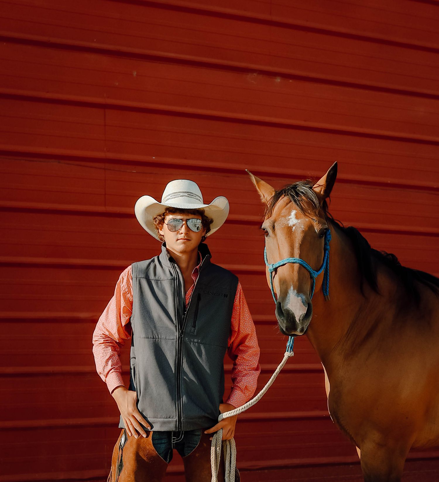 A Curtis, Nebraska lifestyle senior session boy in western attire stands next to a brown horse against a rustic red barn wall.