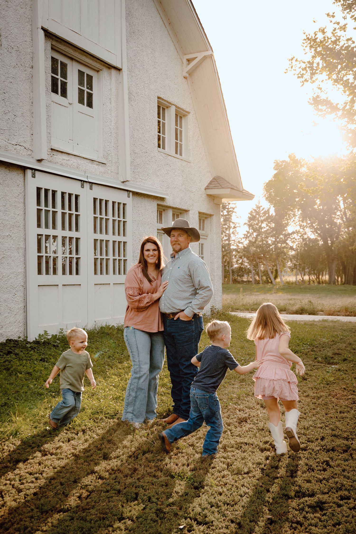 A North Platte, Nebraska lifestyle family session family enjoys a golden sunset moment outside a white barn as children play in the grass.