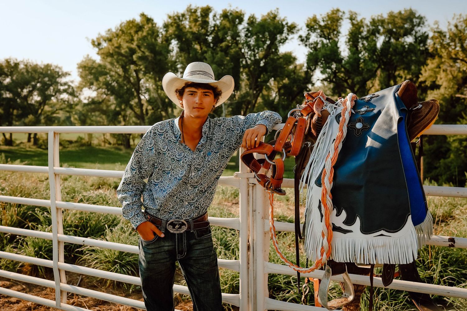 A Curtis, Nebraska lifestyle senior session cowboy in western attire leans against a white ranch fence during golden hour.