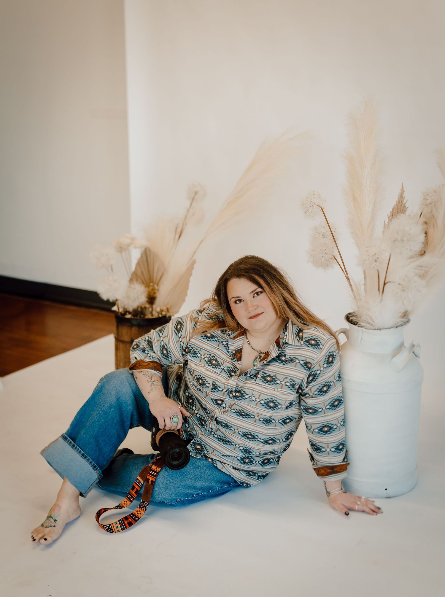 A North Platte, Nebraska lifestyle photographer in a patterned shirt sits on a light colored floor next to pampas grass decor.