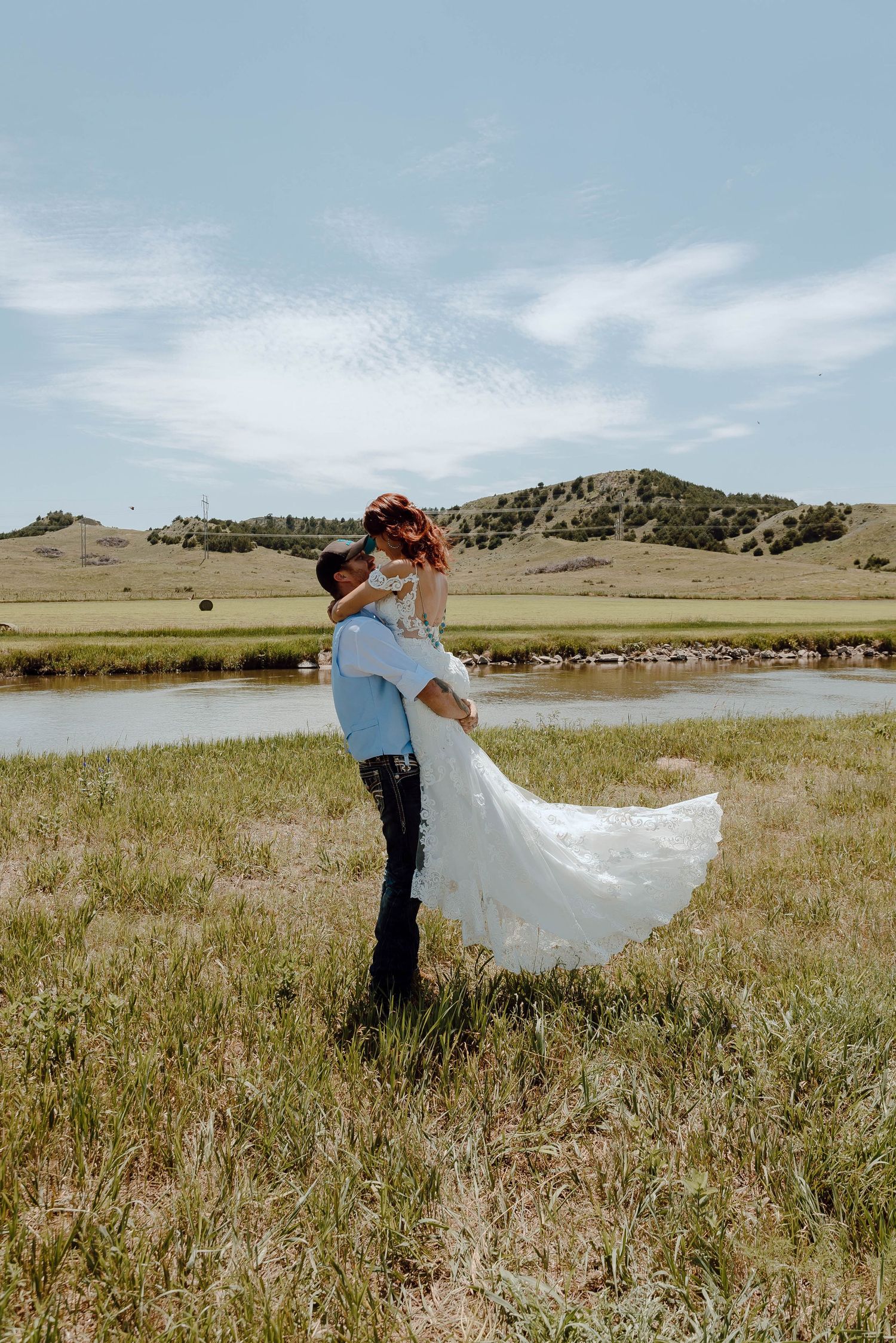 A North Platte, Nebraska lifestyle session couple embraces in a scenic meadow with rolling hills in the background on their wedding day.