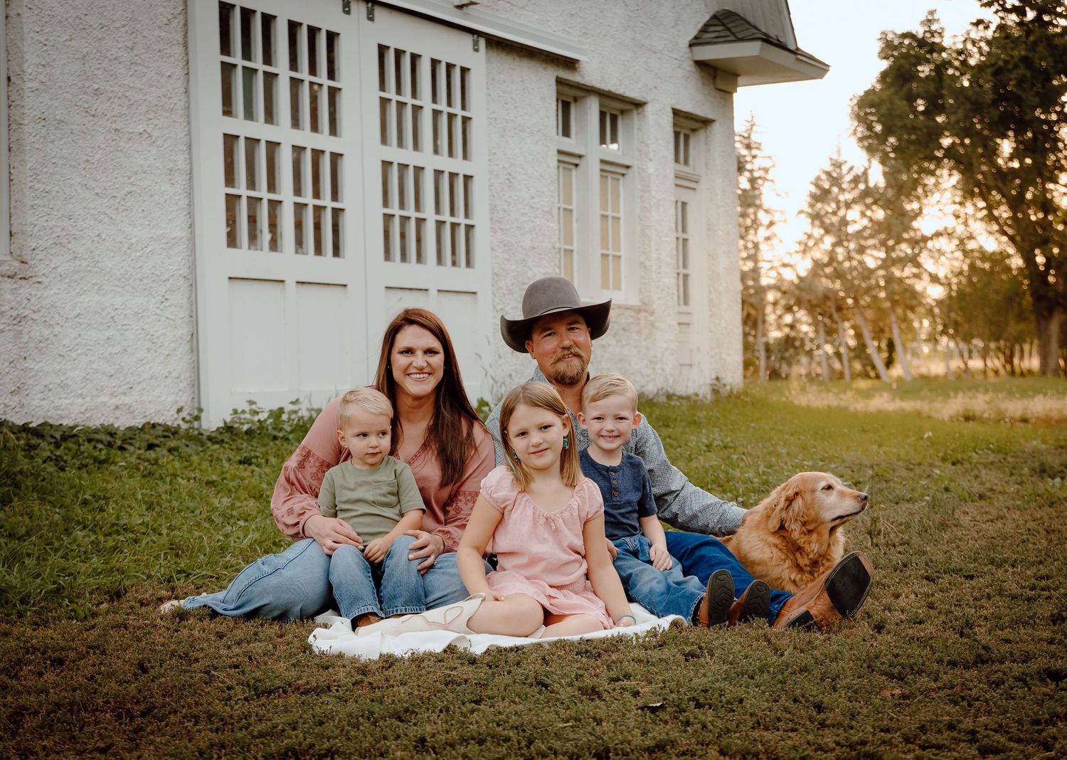 A North Platte, Nebraska lifestyle family session family sits together on the grass outside a white barn during golden hour.