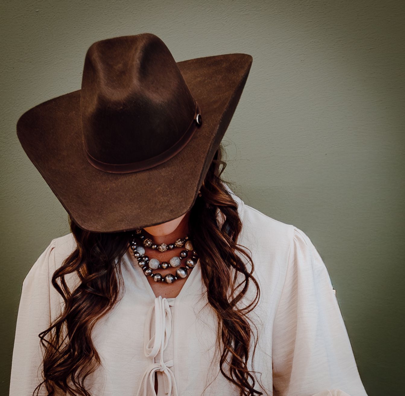 A North Platte, Nebraska lifestyle brand session woman in a brown cowboy hat models jewelry in a close-up shot against a green background.