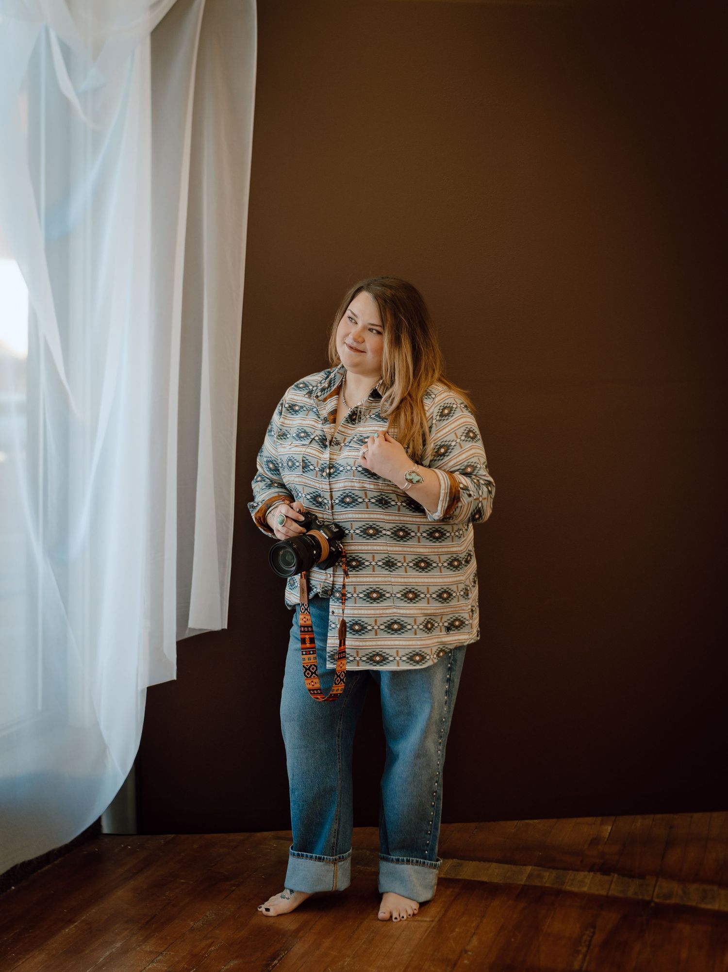 A North Platte, Nebraska lifestyle photographer in a patterned shirt and jeans stands in natural light against a brown background.
