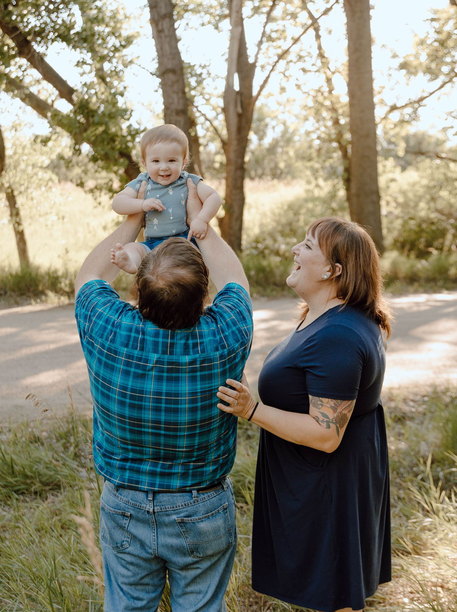 A Sutherland, Nebraska lifestyle family session parents lifting their baby while standing in a sunlit wooded area during golden hour.