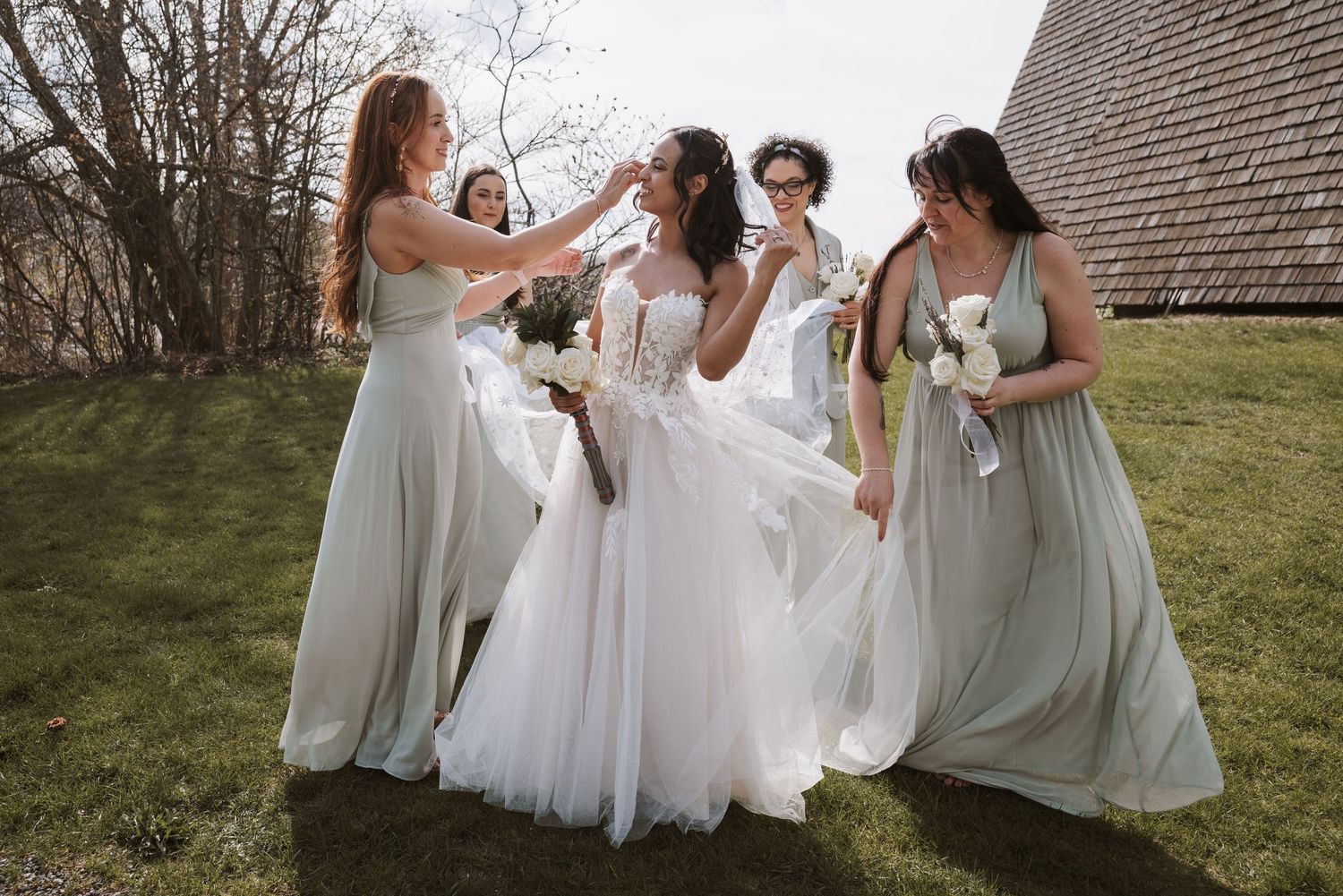 Bride in white gown laughs with bridesmaids in sage green dresses at outdoor wedding.