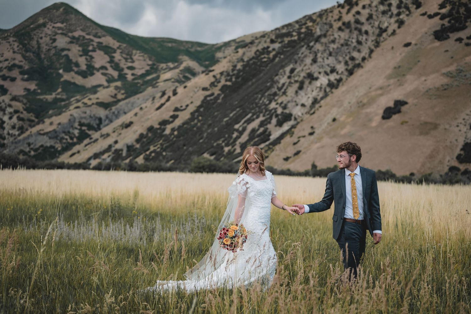 Bride and groom holding hands in a golden field with scenic mountain backdrop on their wedding day.