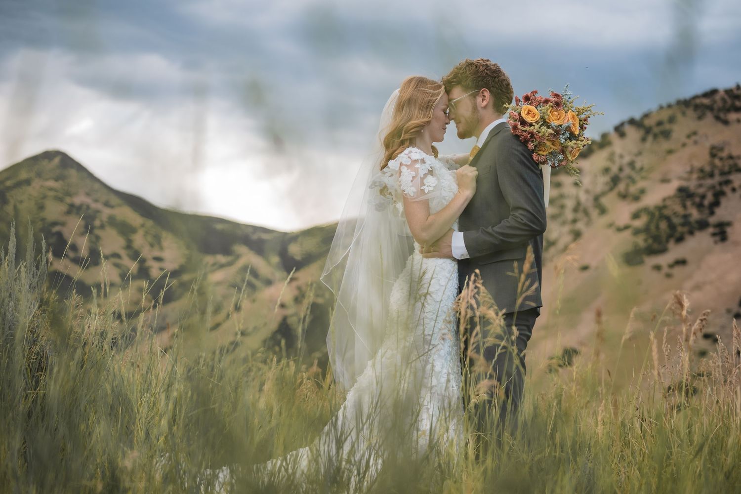 Bride and groom embrace in tall green meadow grass with mountain backdrop.