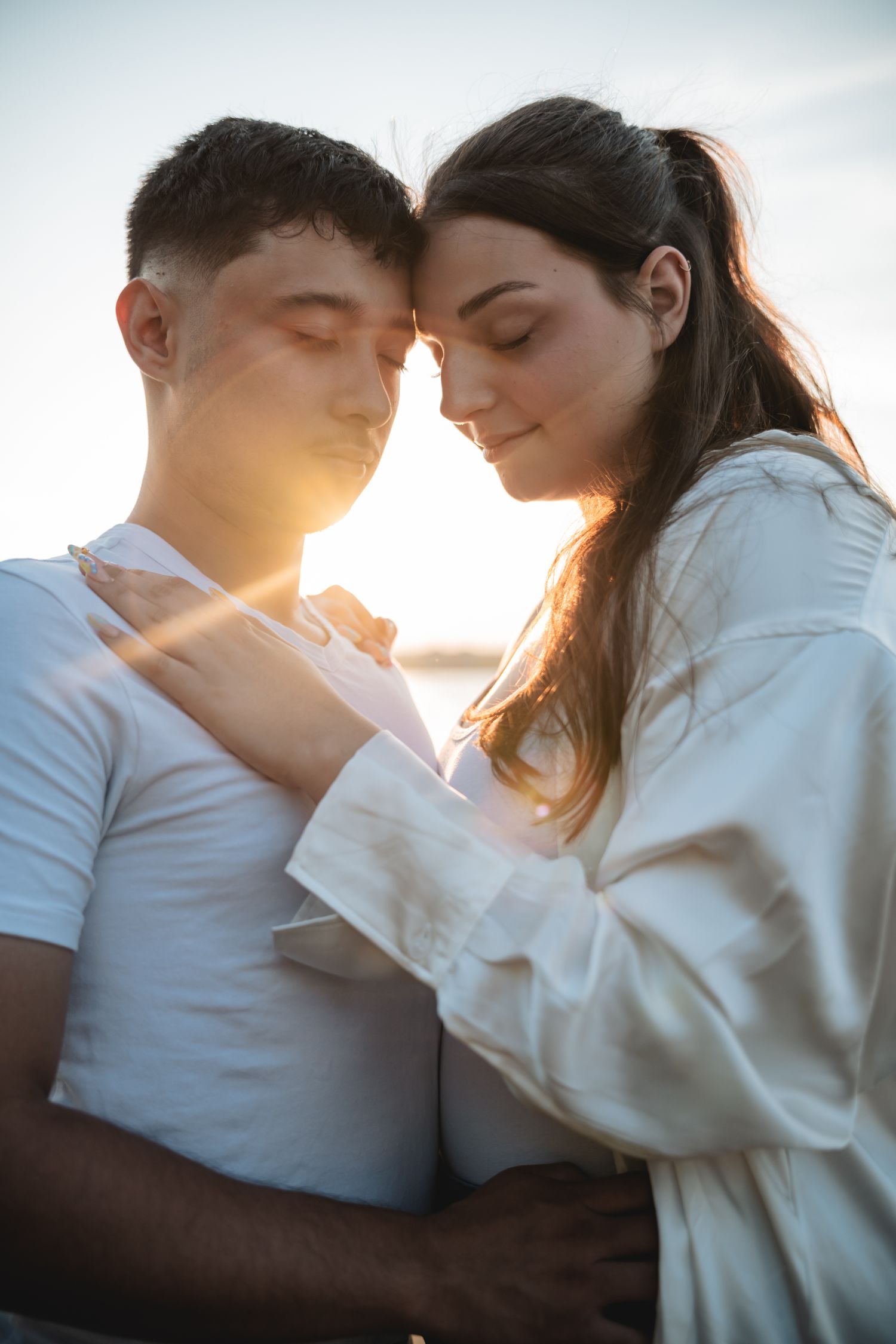Romantic couple embracing at golden hour sunset, woman in white shirt resting head against man in white t-shirt.