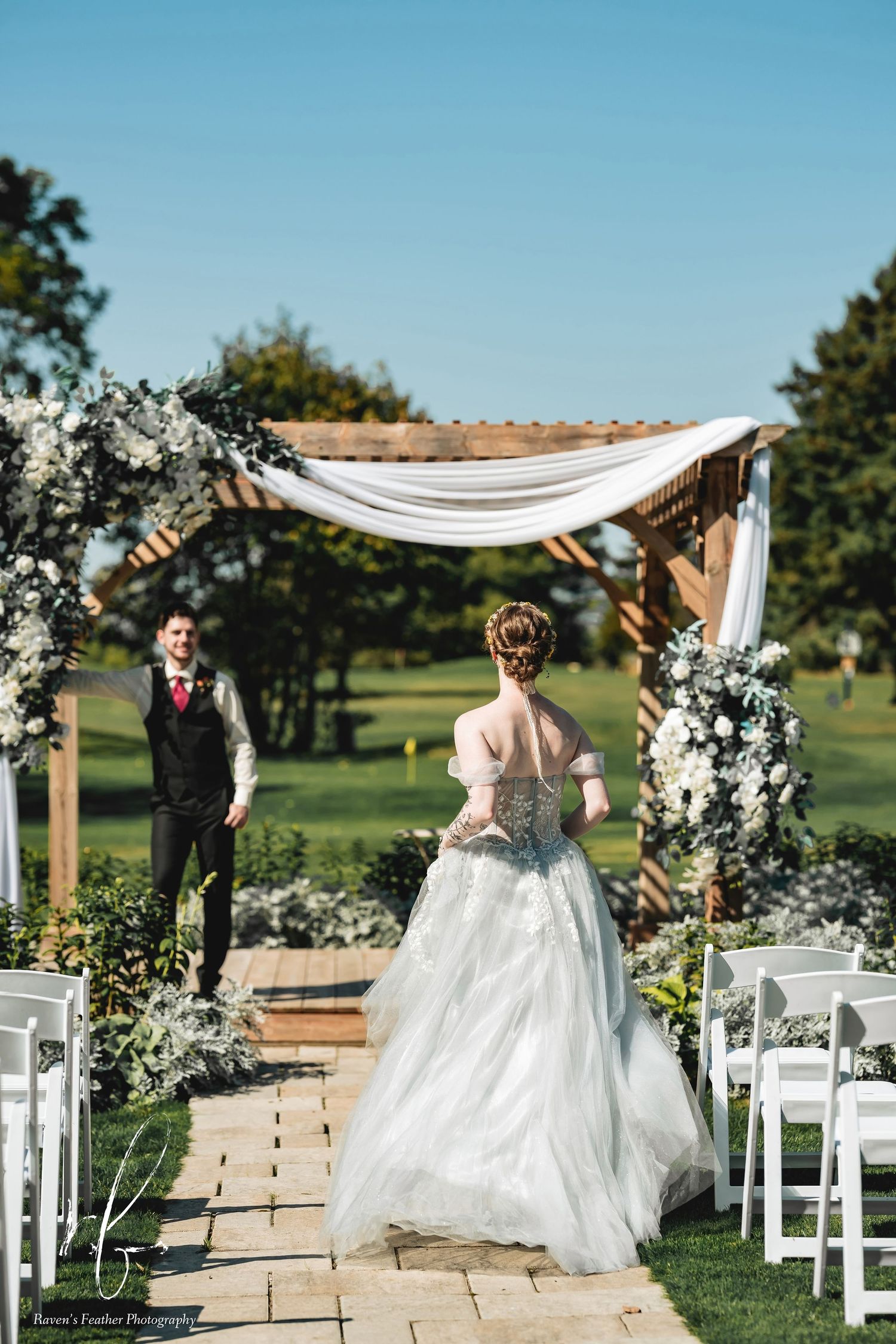 Bride in a flowing gown walks toward groom at an outdoor wedding ceremony beneath a floral wooden arch.