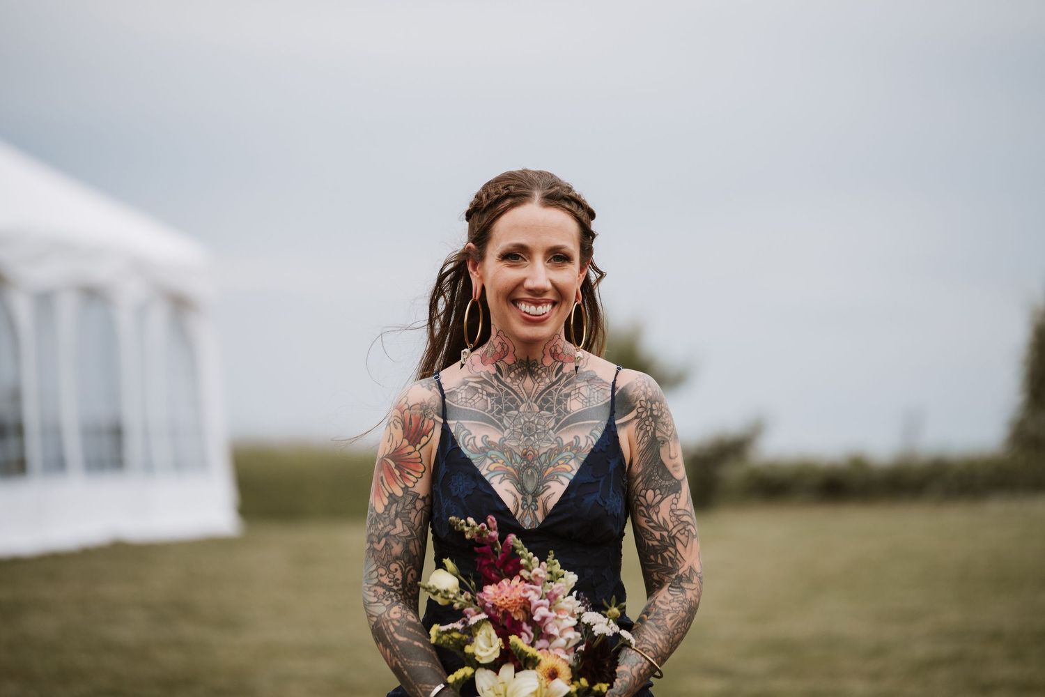 Tattooed bridesmaid smiles holding colorful bouquet at outdoor wedding venue.