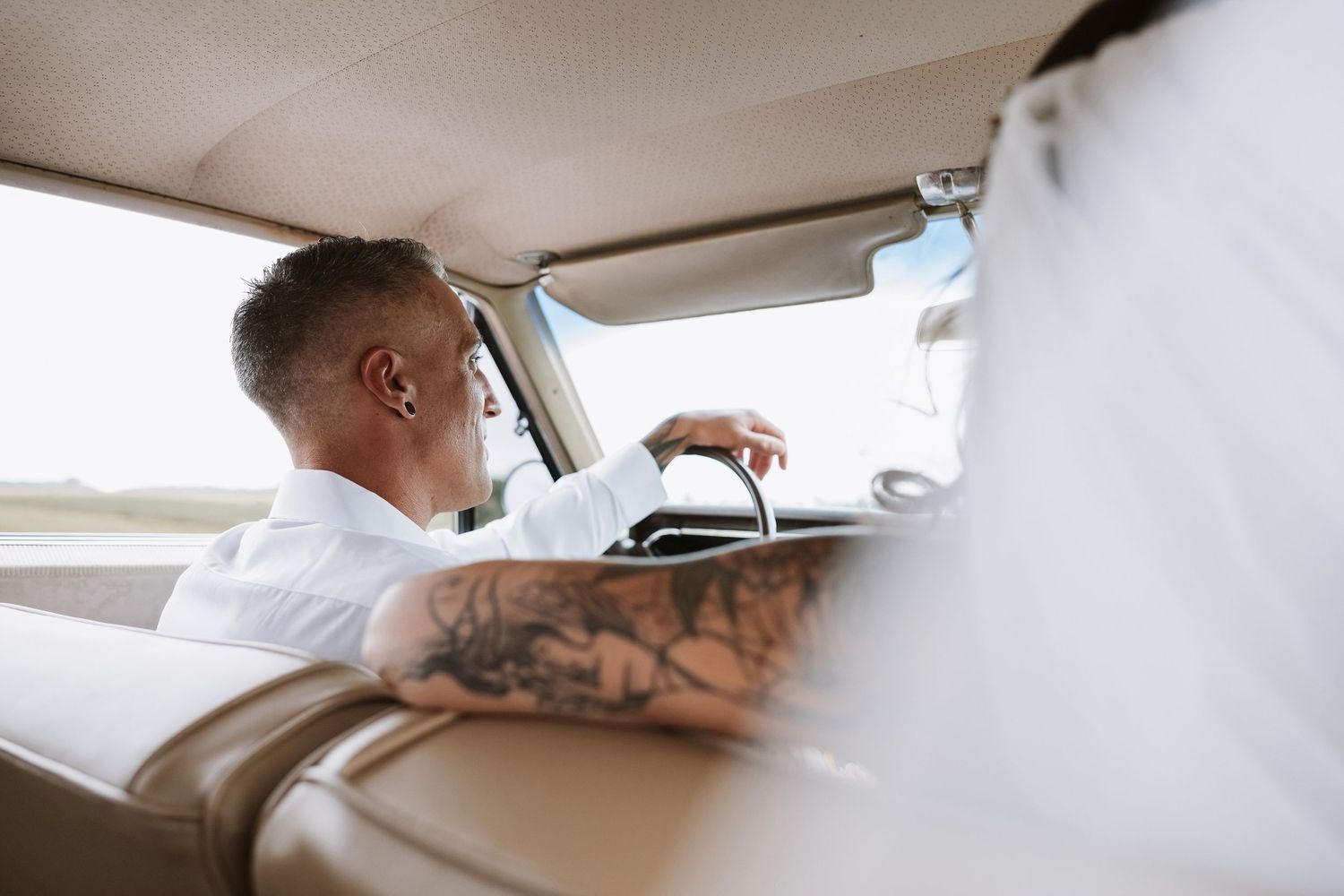 Tattooed groom in white shirt driving a vintage car on his wedding day.