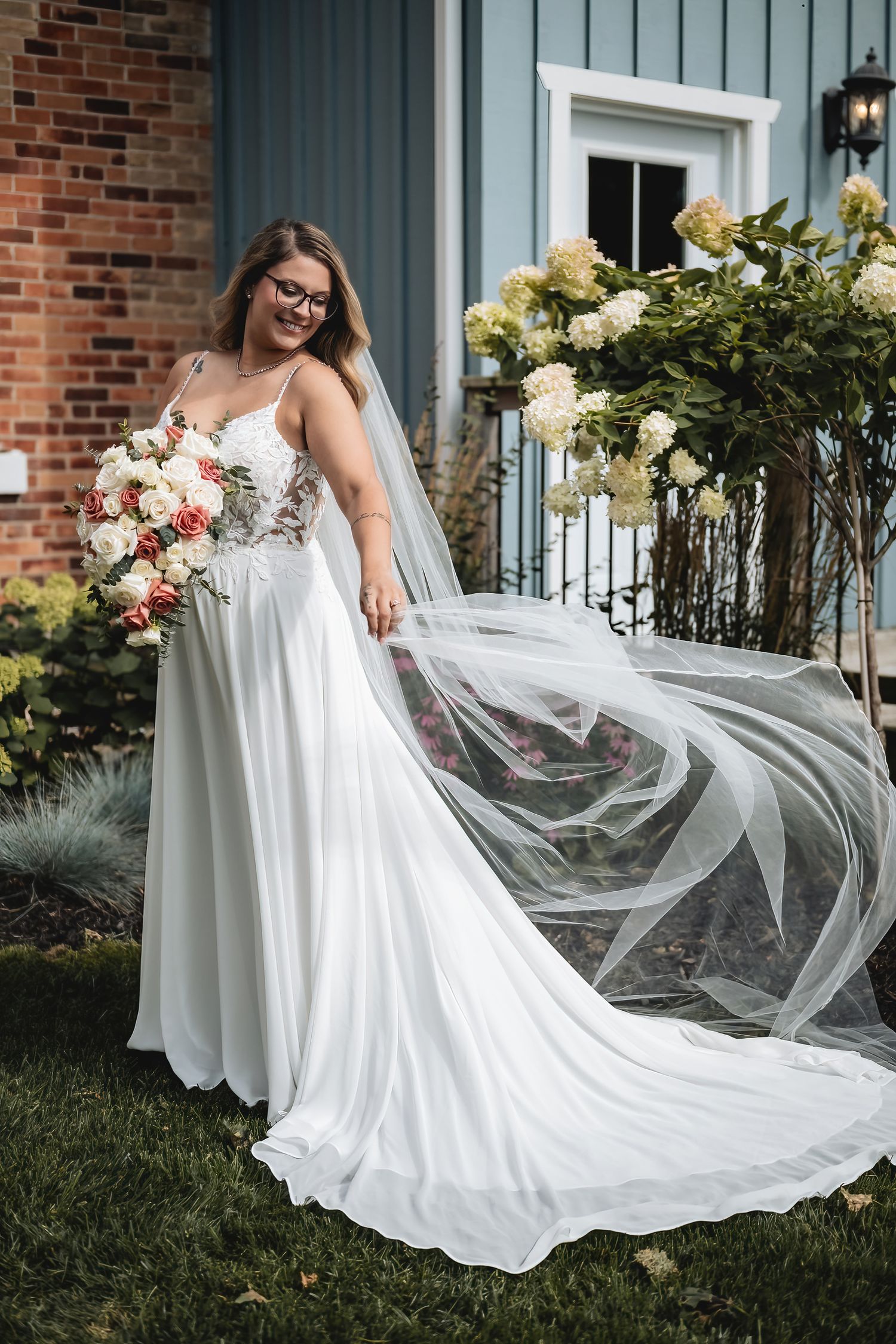 Smiling bride in white lace A-line gown with flowing veil holds bouquet outdoors near hydrangeas.