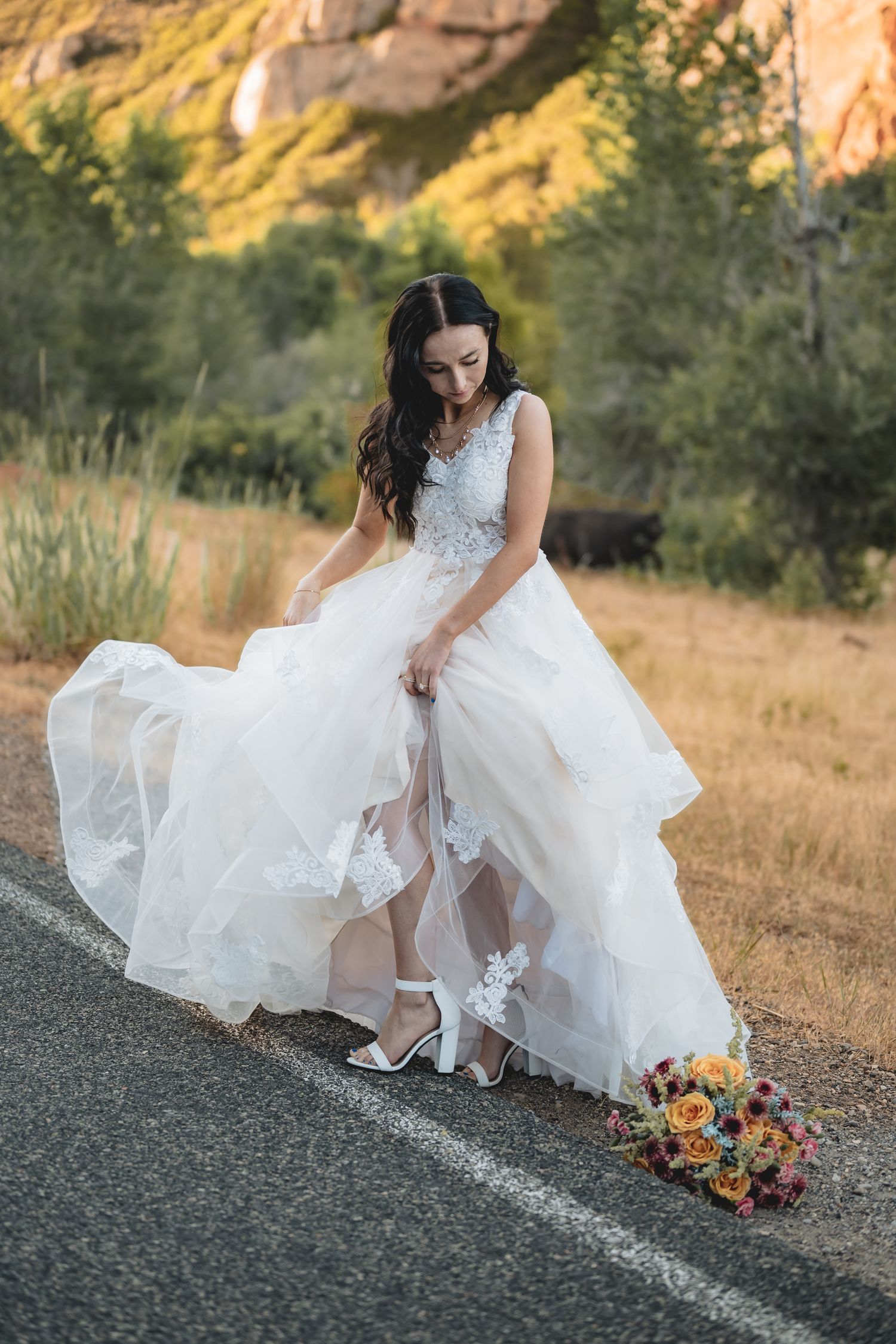 Bride in layered tulle wedding gown adjusting her shoe on a scenic mountain road.