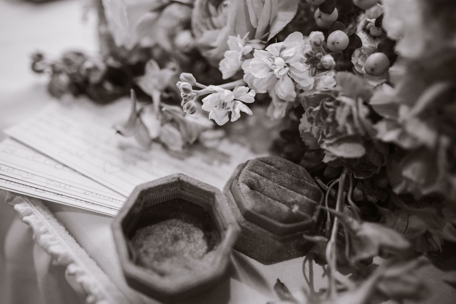 Black and white flat lay of wedding rings in wooden box with floral bouquet and stationery.