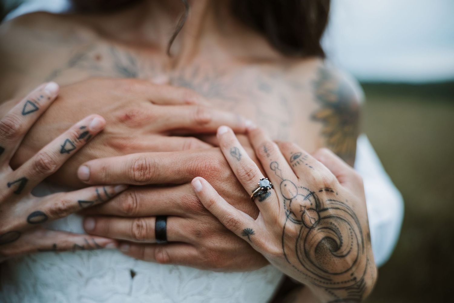 Tattooed couple holding hands on brides chest showing wedding rings, intricate ink designs on fingers and arms.