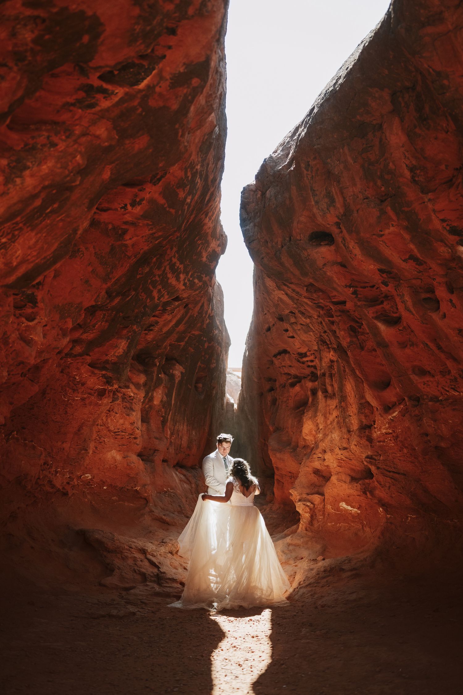 Bride and groom stand together in a narrow red rock canyon with light streaming from above.