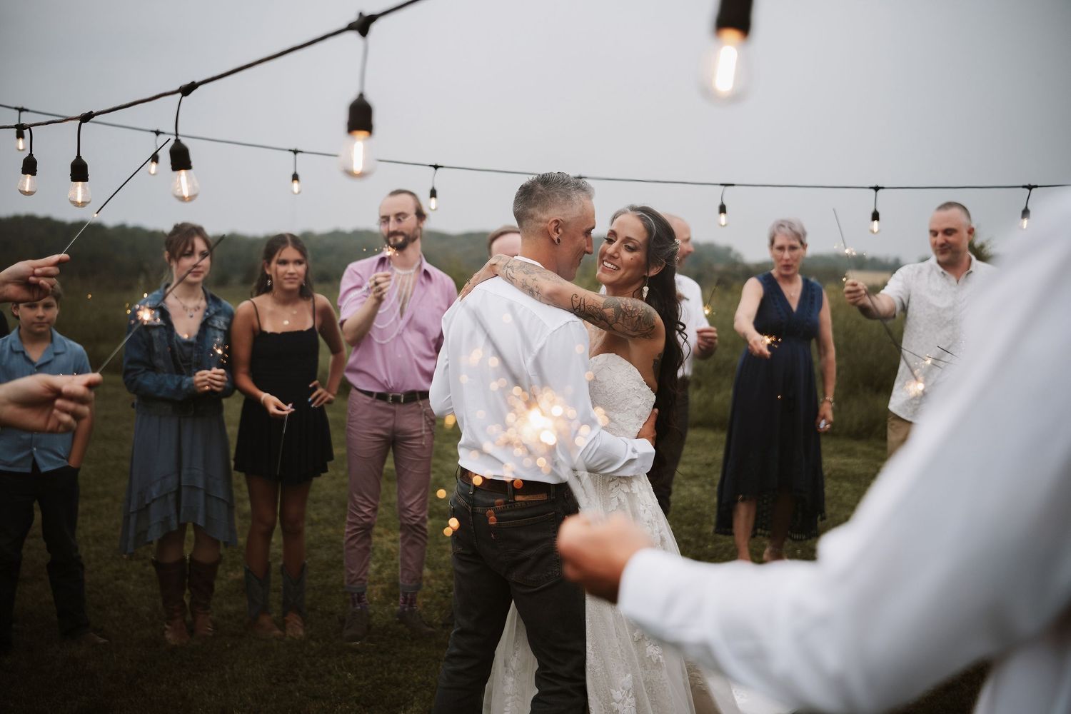 Couple shares first dance surrounded by guests holding sparklers under string lights at outdoor wedding reception.