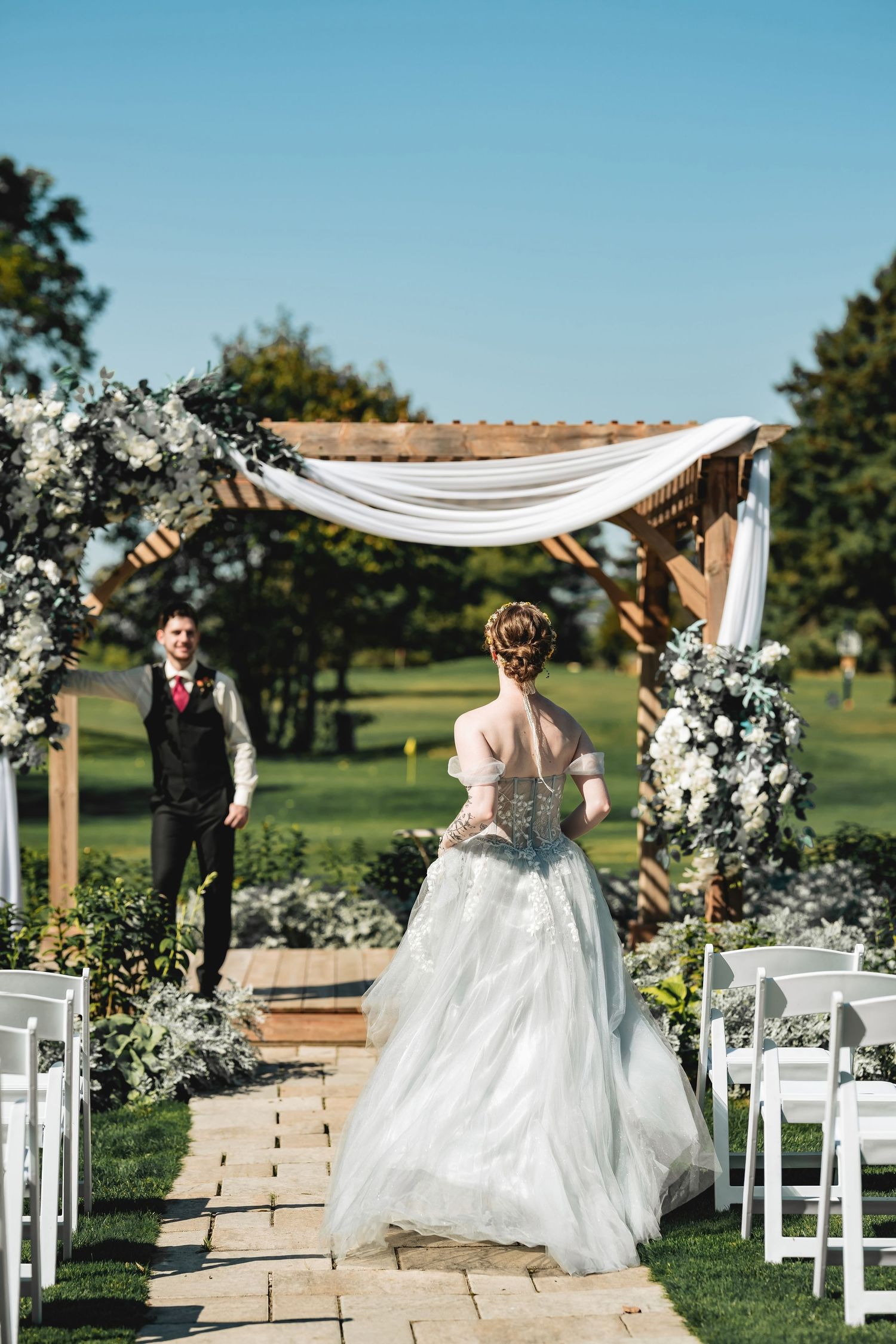 Bride in blue-grey ballgown walking toward groom at floral outdoor wedding ceremony altar.