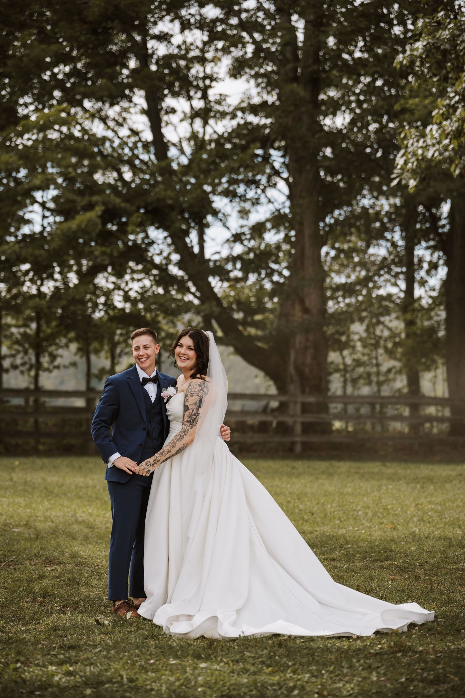 Bride and groom pose together on lush green lawn with large trees and wooden fence in background on wedding day.