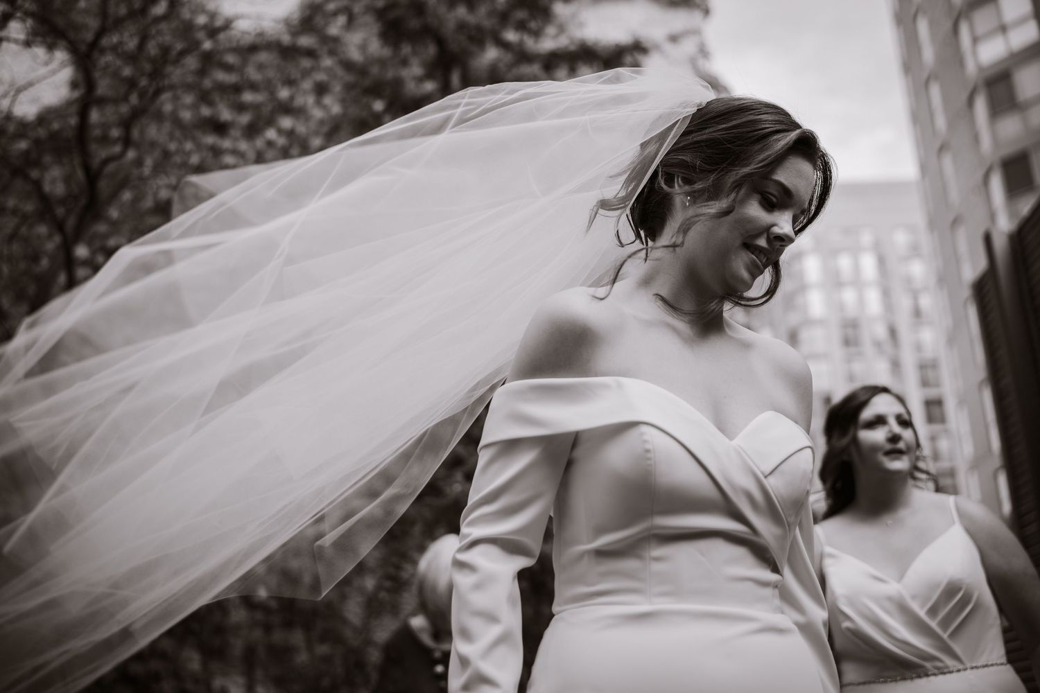 Black and white photo of radiant bride with veil blowing in wind on city street.