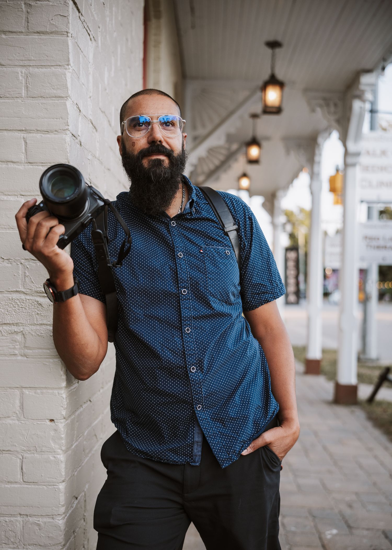 Bearded male photographer with glasses holding a camera, leaning against a white brick wall on a covered sidewalk.