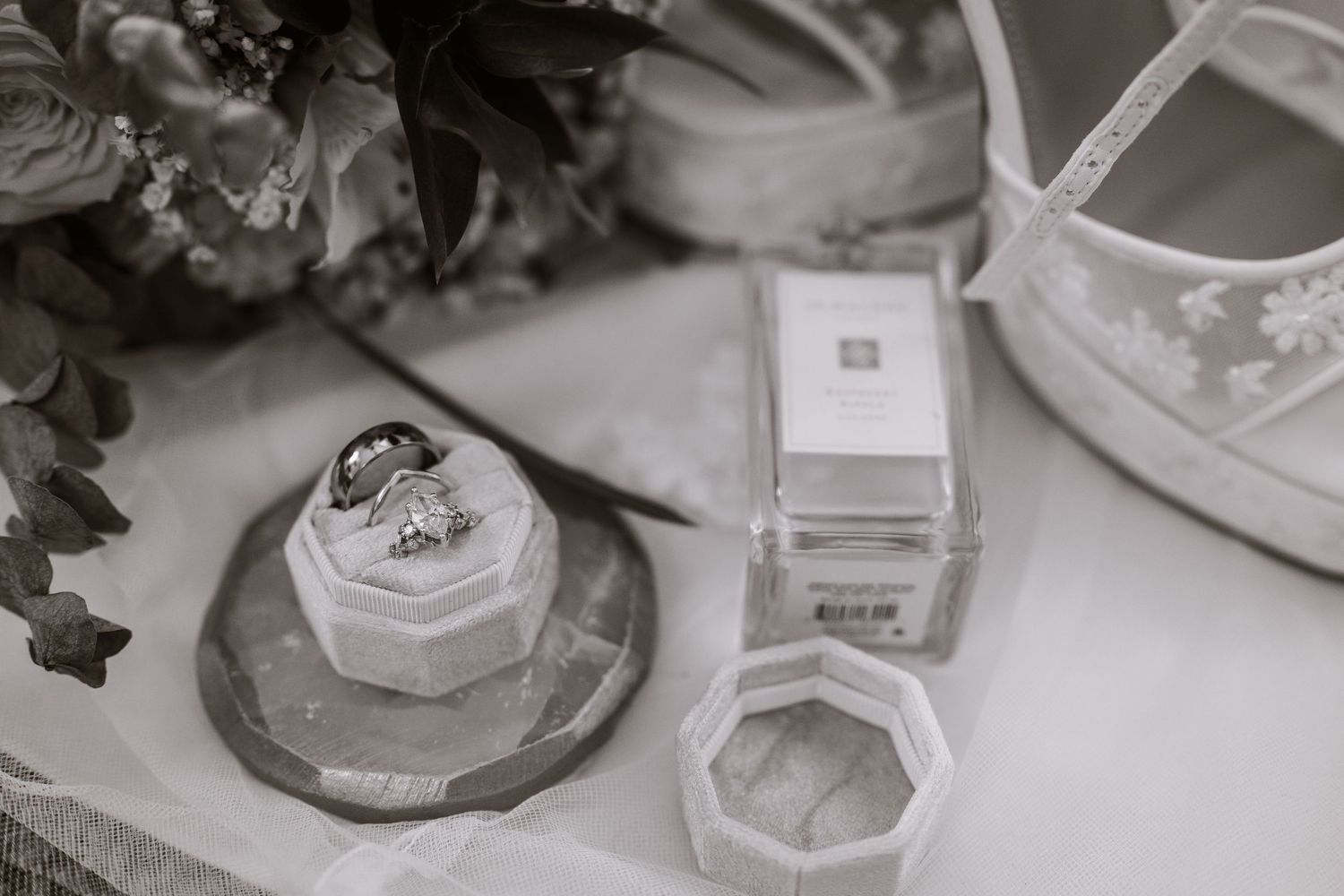 Black and white wedding flat lay with diamond rings on velvet holder, Jo Malone perfume box, and lace bridal shoes.