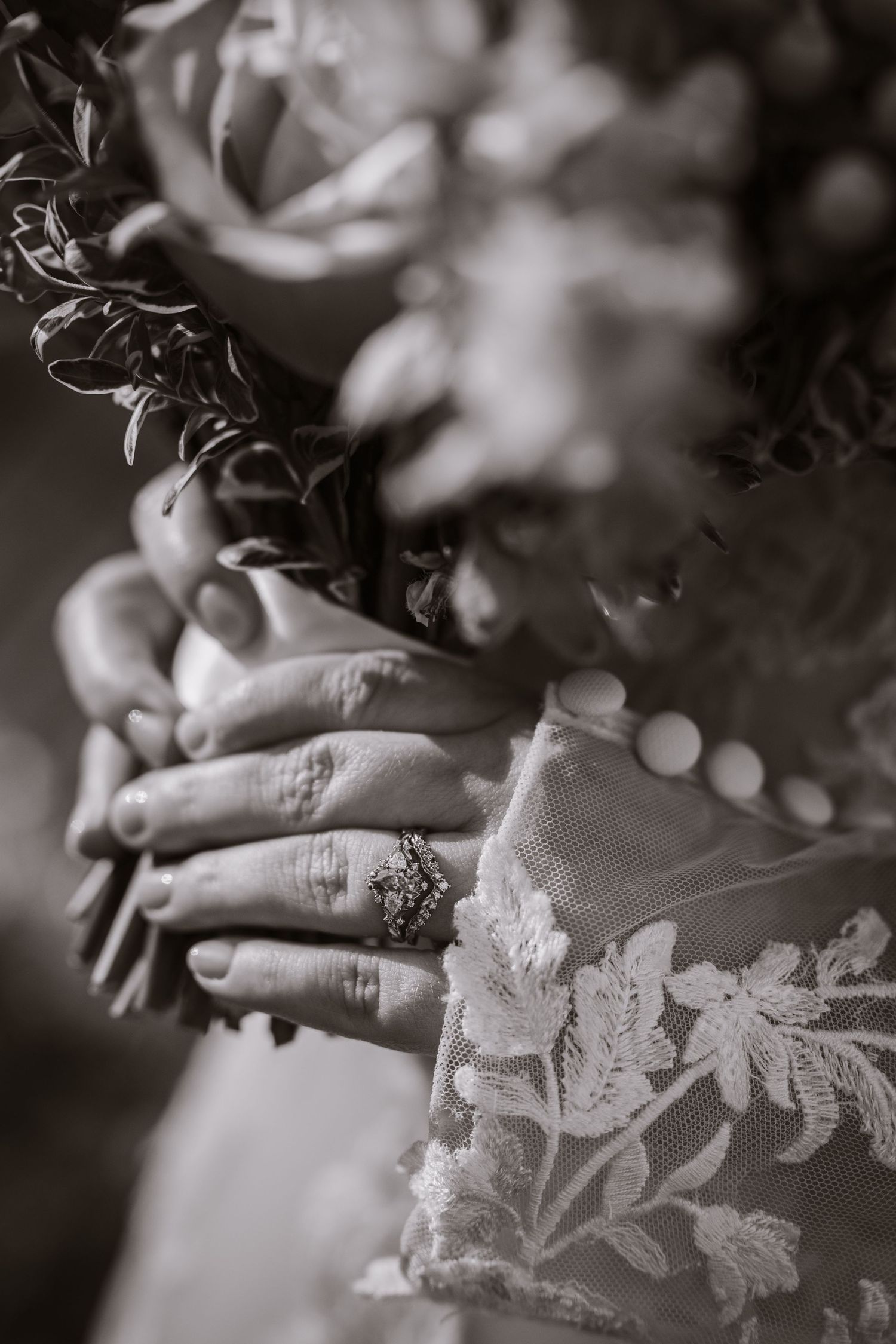 Black and white close-up of bride holding floral bouquet wearing lace gown and ornate ring.