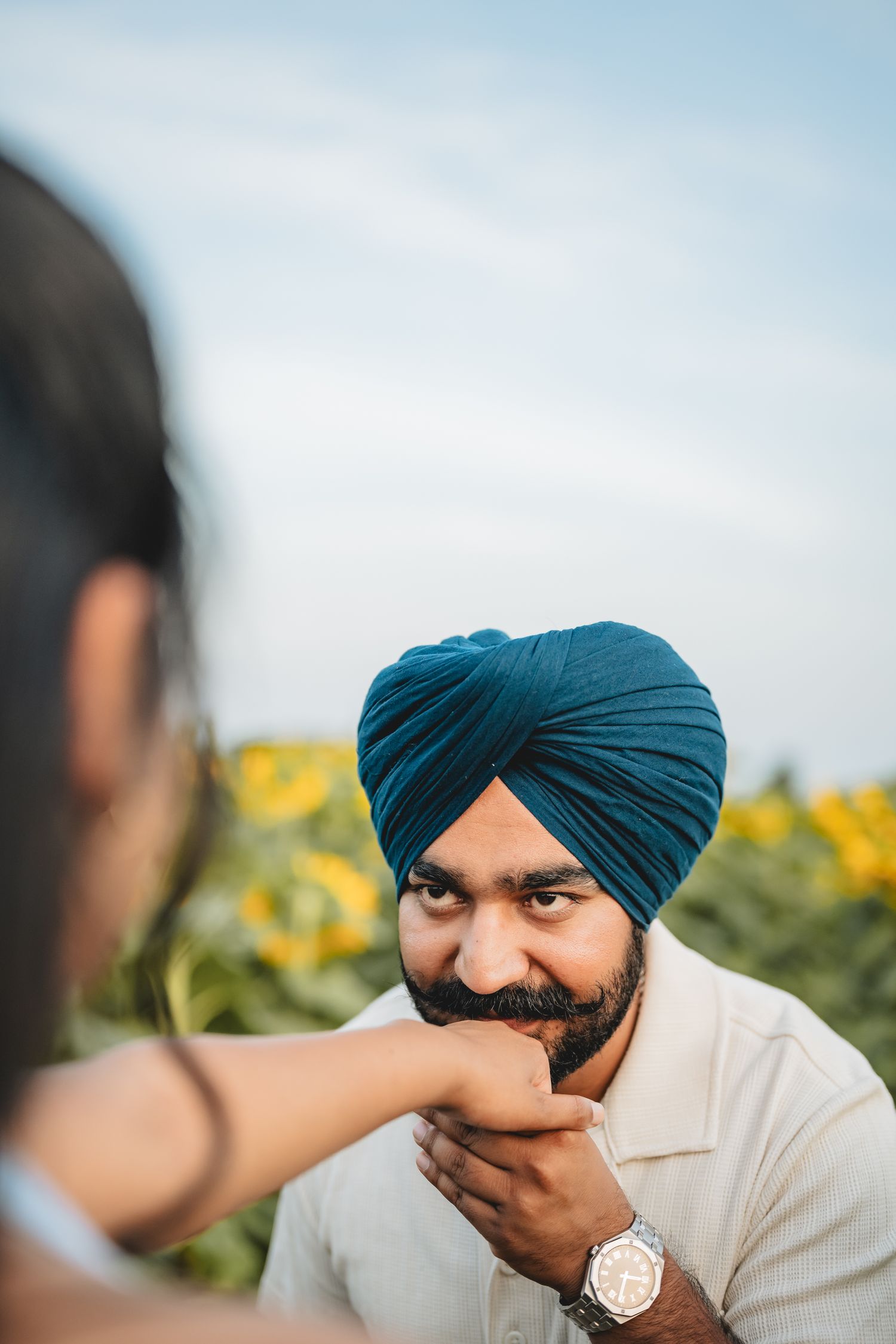A Sikh man wearing a blue turban and watch smiles as a woman gently holds his face near a sunflower field.