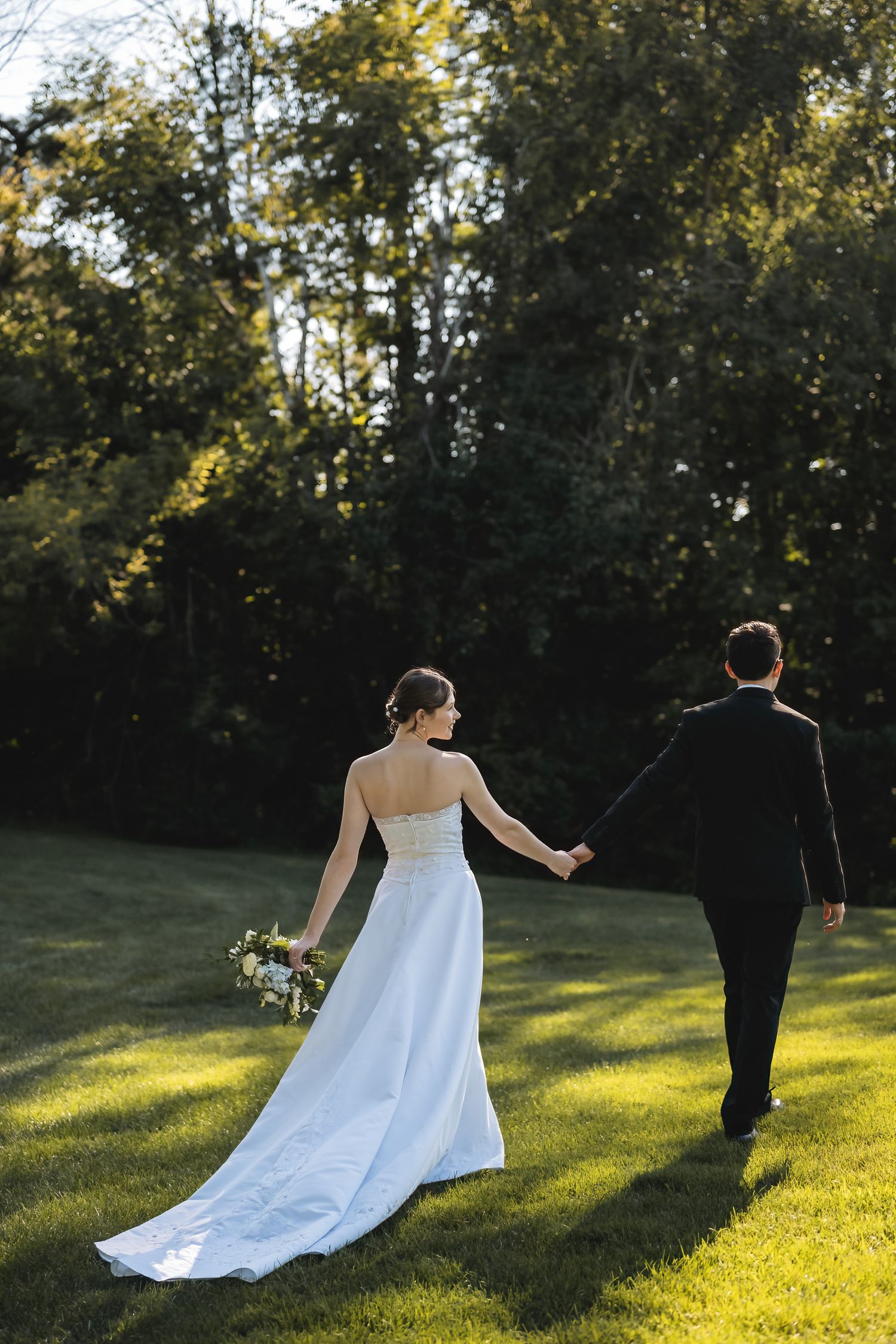 Bride and groom walking hand in hand across lush green lawn surrounded by trees on their wedding day.