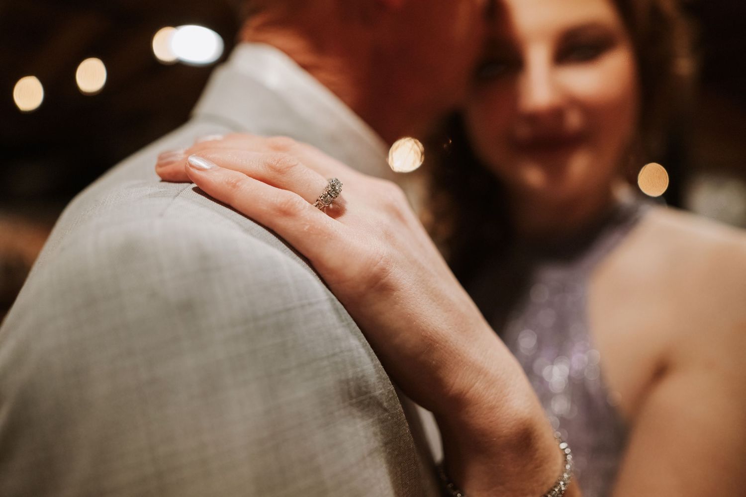 Close-up of woman's hand with diamond ring resting on man's gray suit jacket at wedding reception.