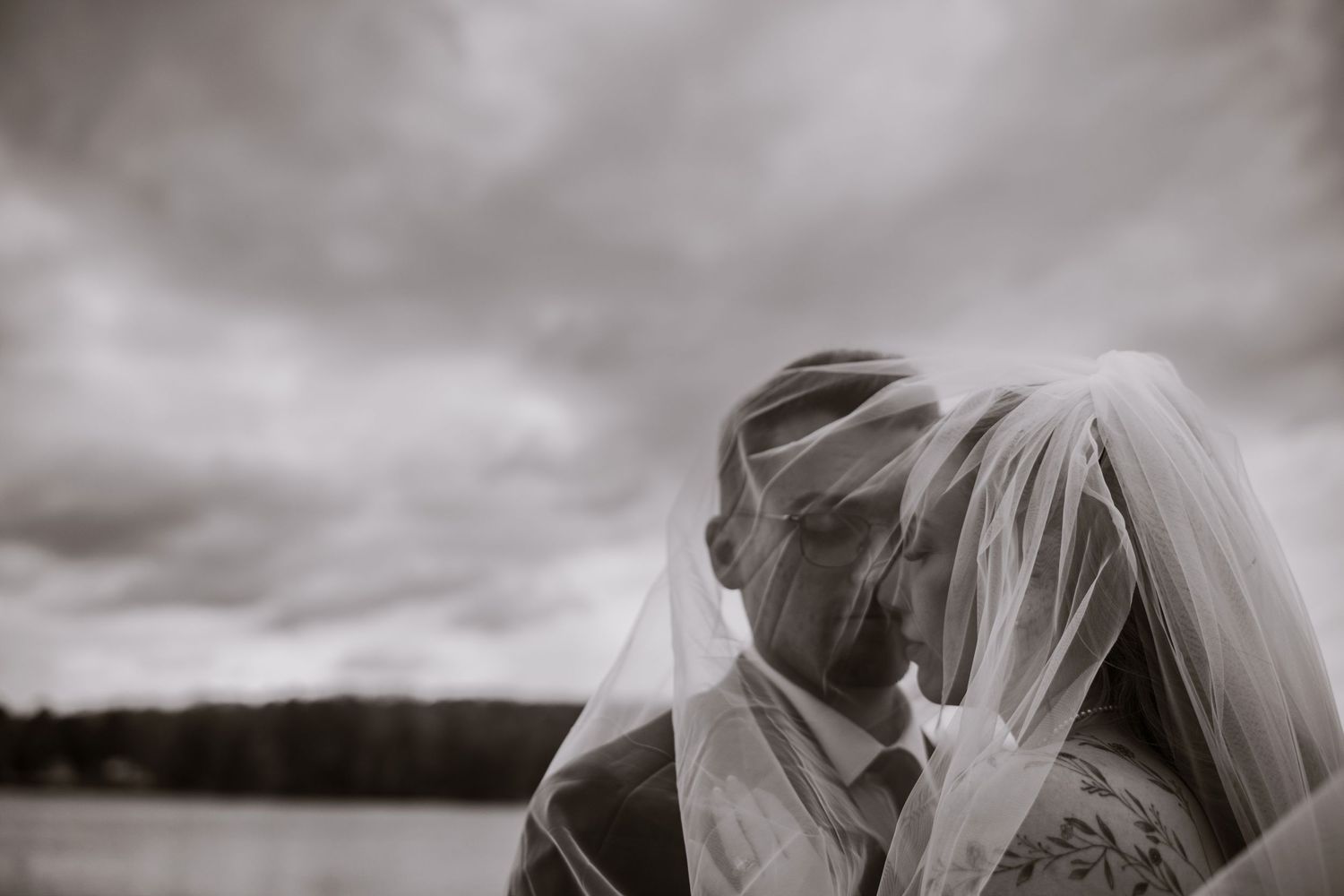 Bride and groom share intimate moment under flowing veil near lake on cloudy day.