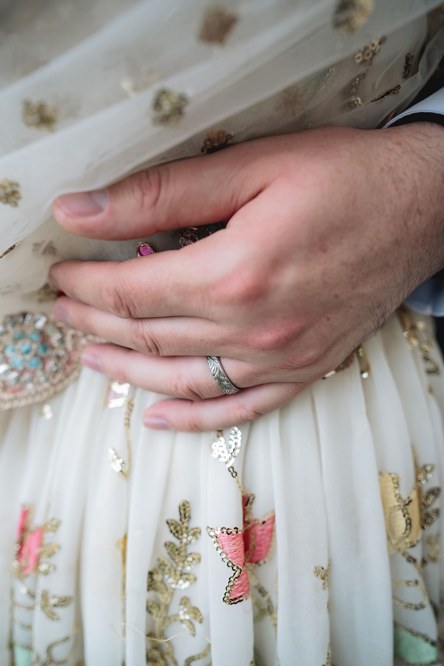 Close-up of couple's hands with wedding ring on embroidered white bridal lehenga with gold and pink floral details.