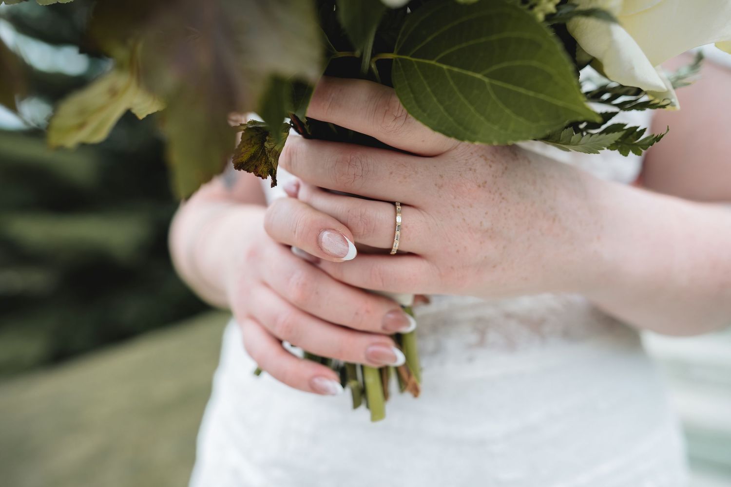 Close-up of bride's hands holding a green leaf bouquet, wearing a gold wedding band with neutral manicure.