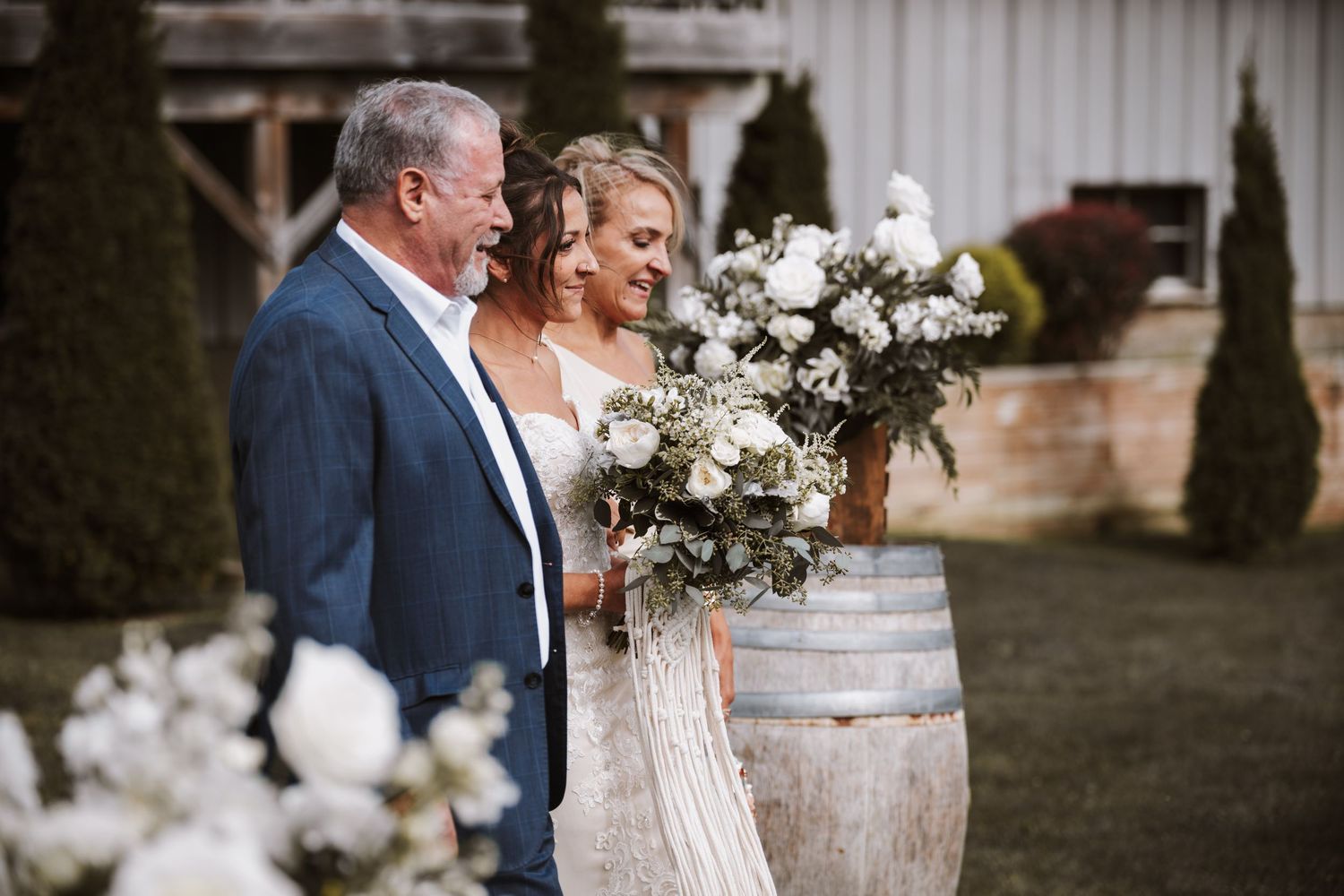 Bride with large white floral bouquet stands beside father & mother at rustic barn wedding.