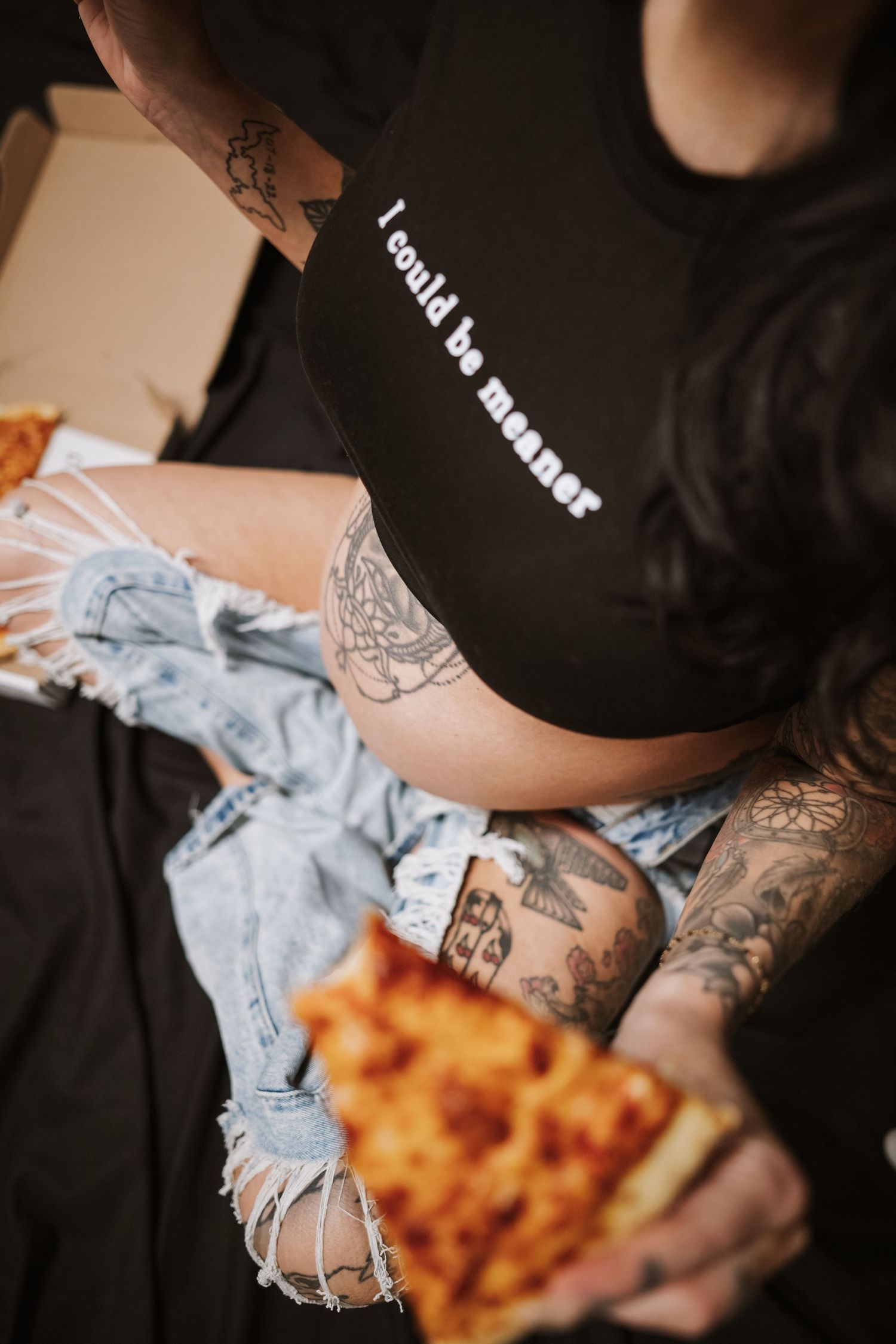 Tattooed woman in black crop top and ripped jeans holding a slice of pizza.