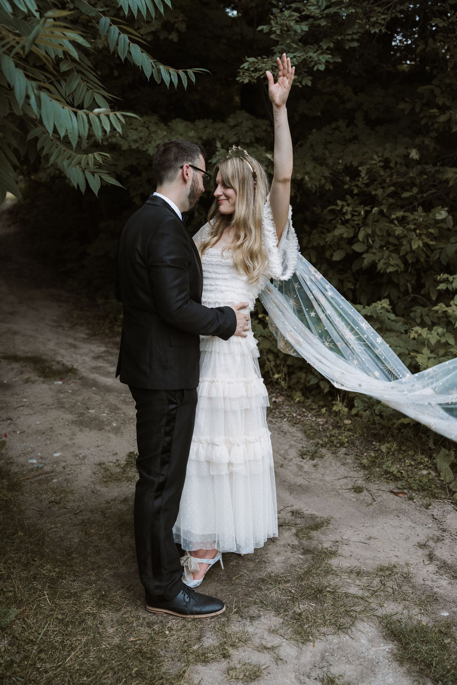 Wedding couple dancing outdoors, bride in flowing white dress with veil raised against green trees.