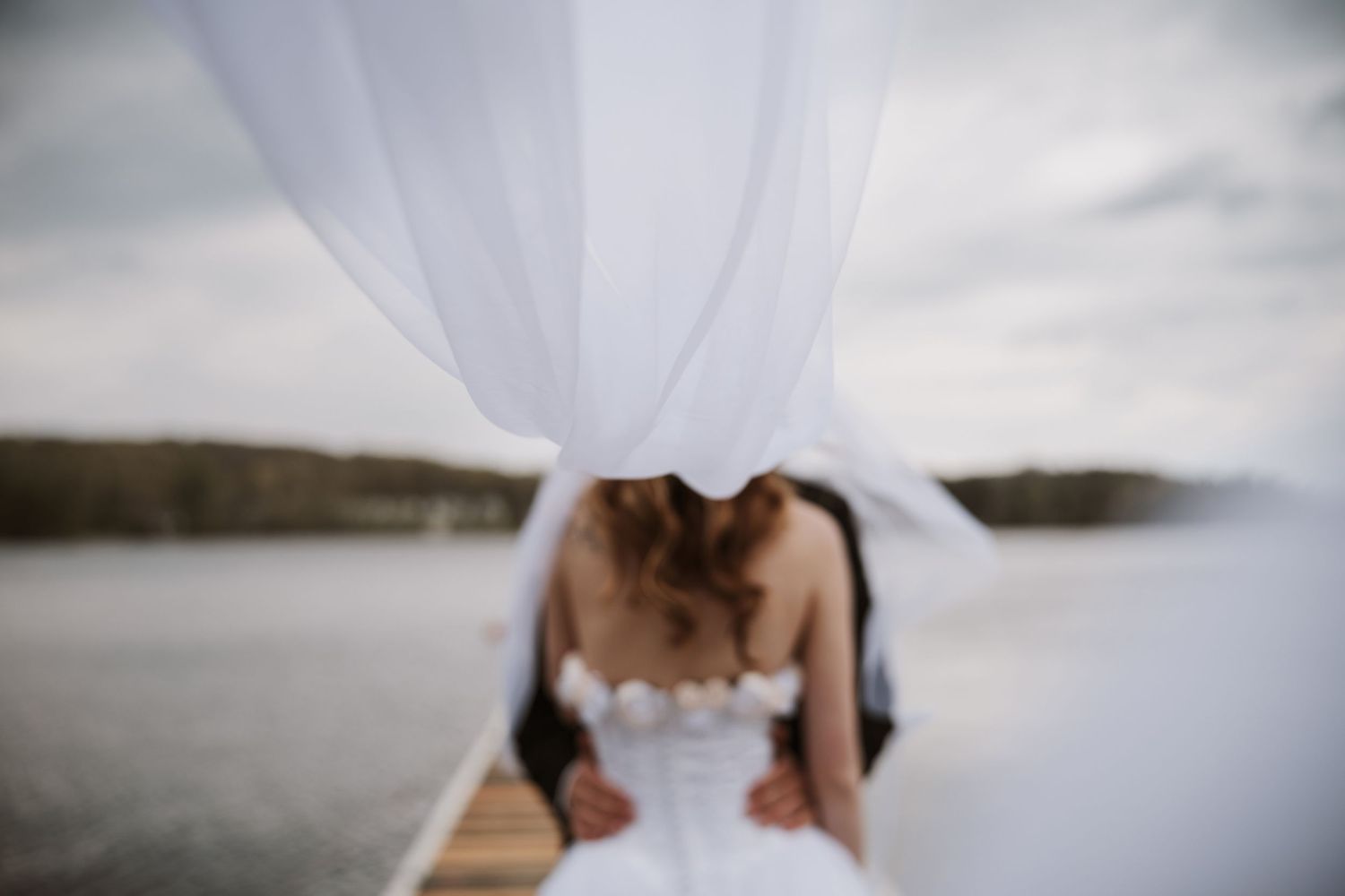 Romantic wedding couple embraces on wooden dock as white veil billows over them.