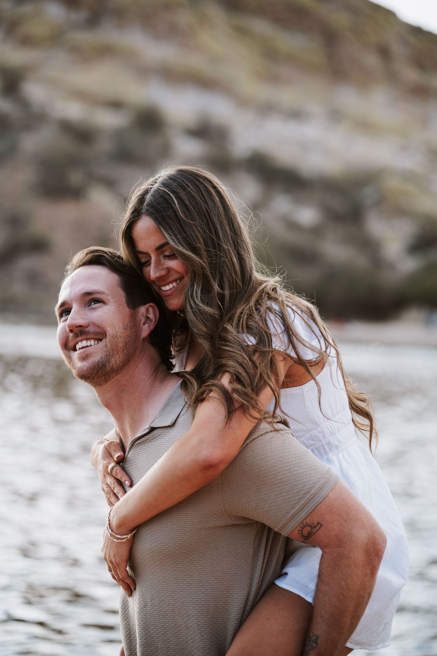 Joyful man gives smiling woman a piggyback ride near a scenic lake with rocky backdrop.