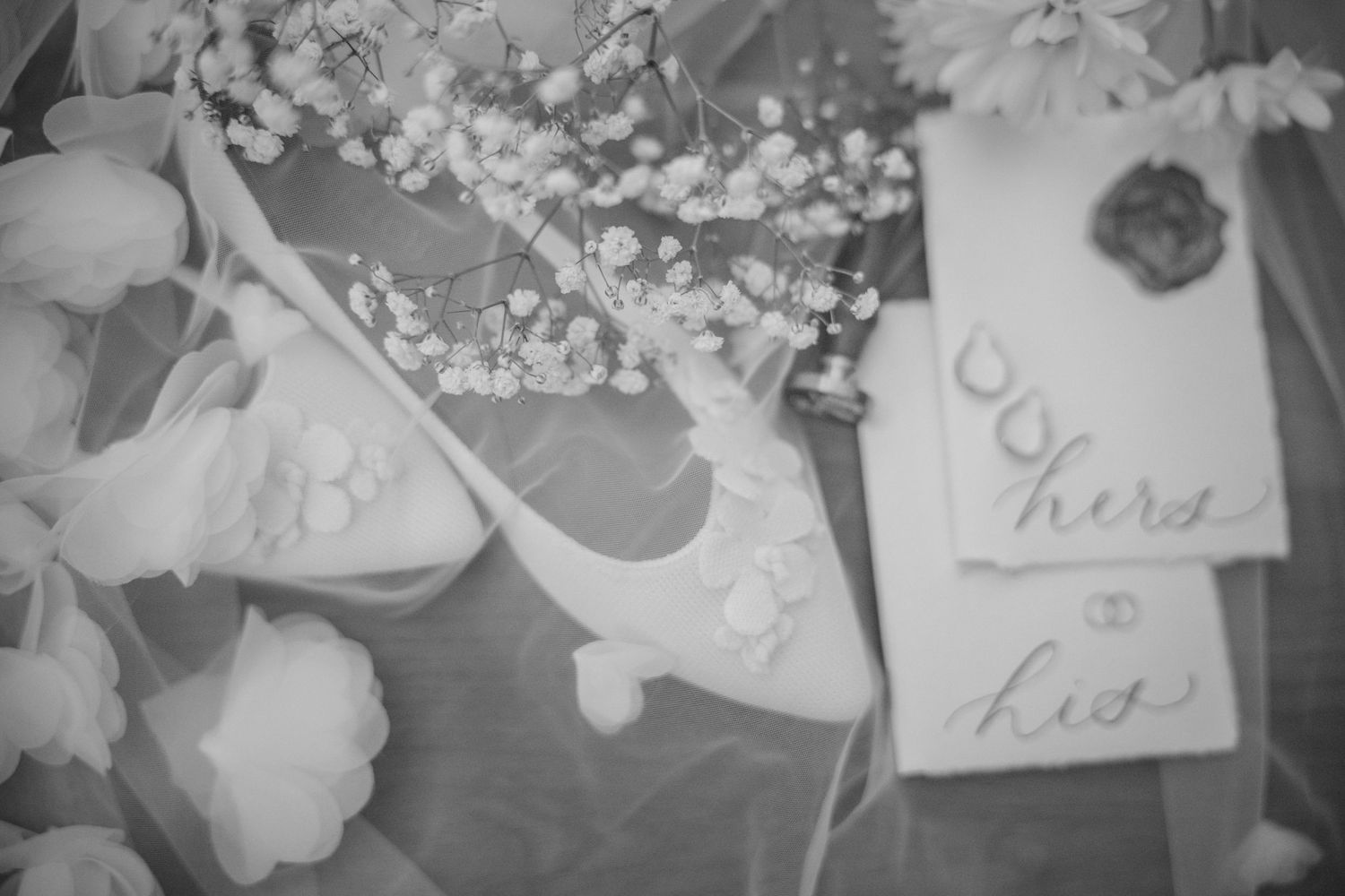 Black and white wedding flat lay with a bridal shoe, baby's breath flowers, and calligraphy vow cards labeled hers and his.