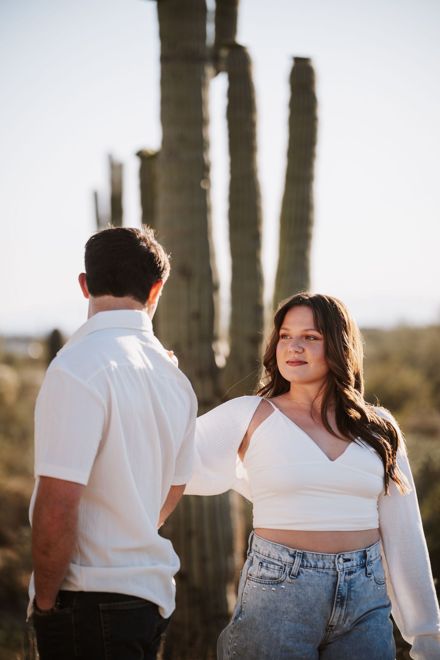 Young woman in white crop top and jeans standing near a saguaro cactus in desert landscape.