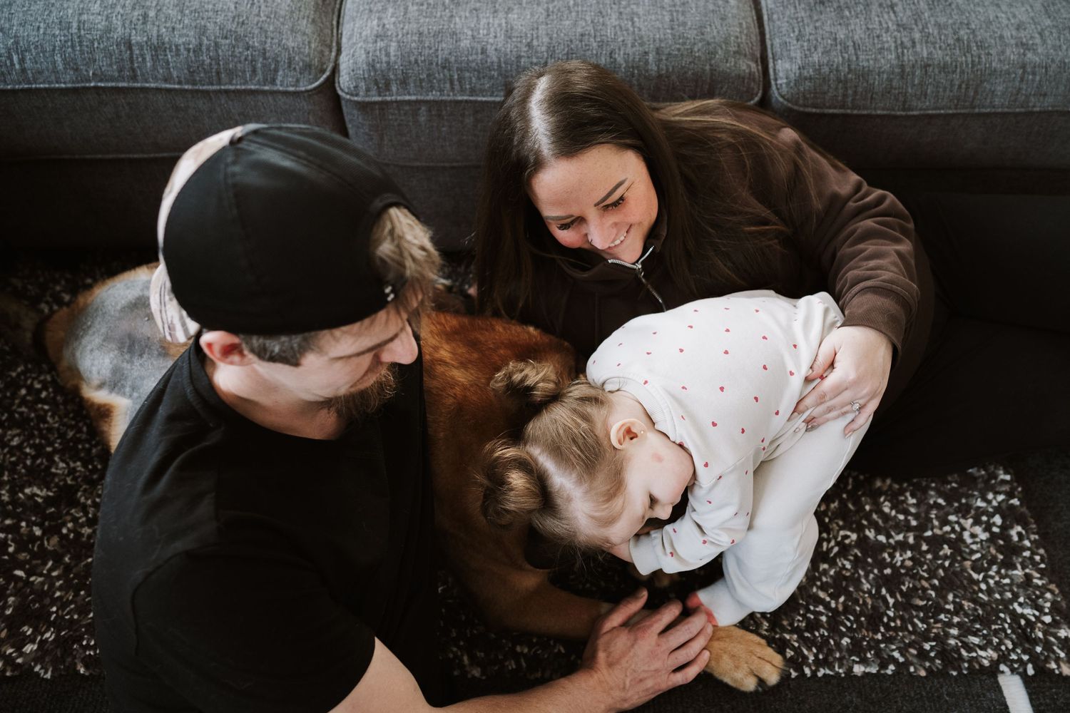 Family with young child and dog relaxing together on floor near gray sofa.