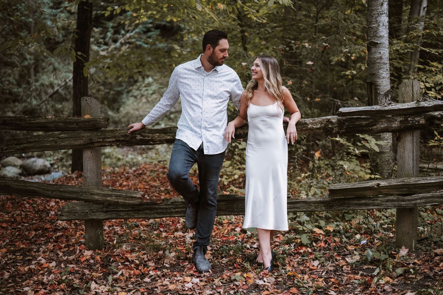 Couple walking together along rustic wooden fence surrounded by autumn foliage in forest setting.