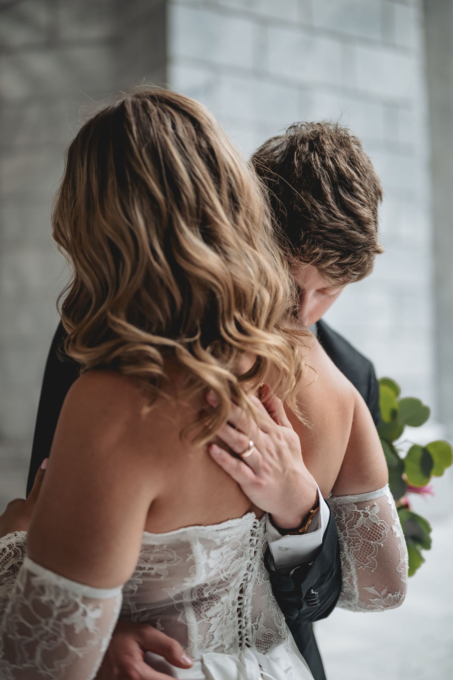 Groom tenderly kisses bride's shoulder as she wears off-shoulder lace wedding gown indoors.