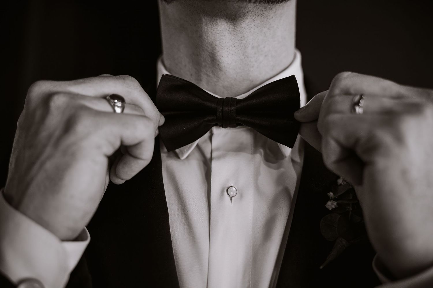 Black and white photo of a groom adjusting his bow tie, wearing a tuxedo with rings on his fingers.
