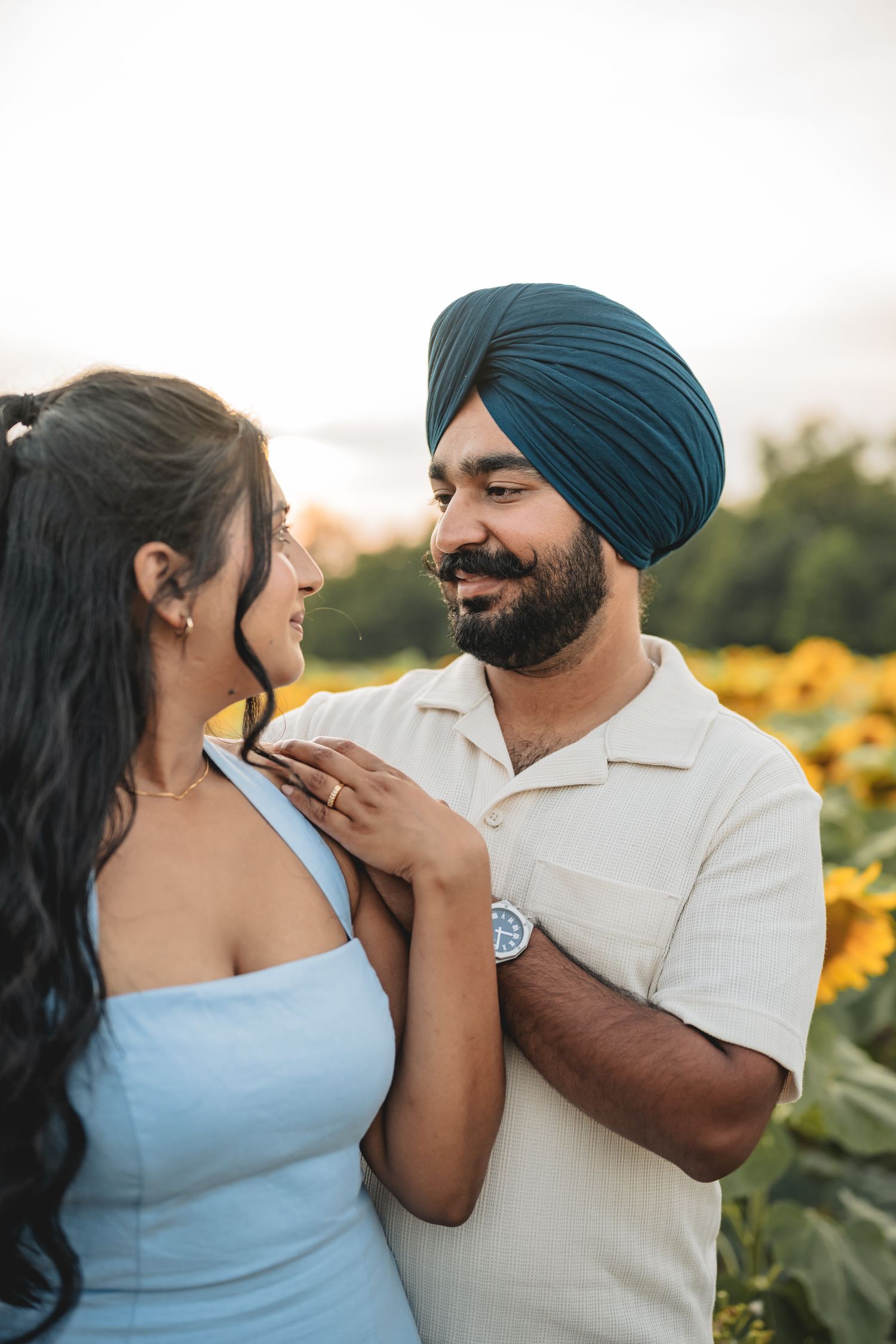 Sikh man in teal turban and woman share tender moment in sunflower field at golden hour.
