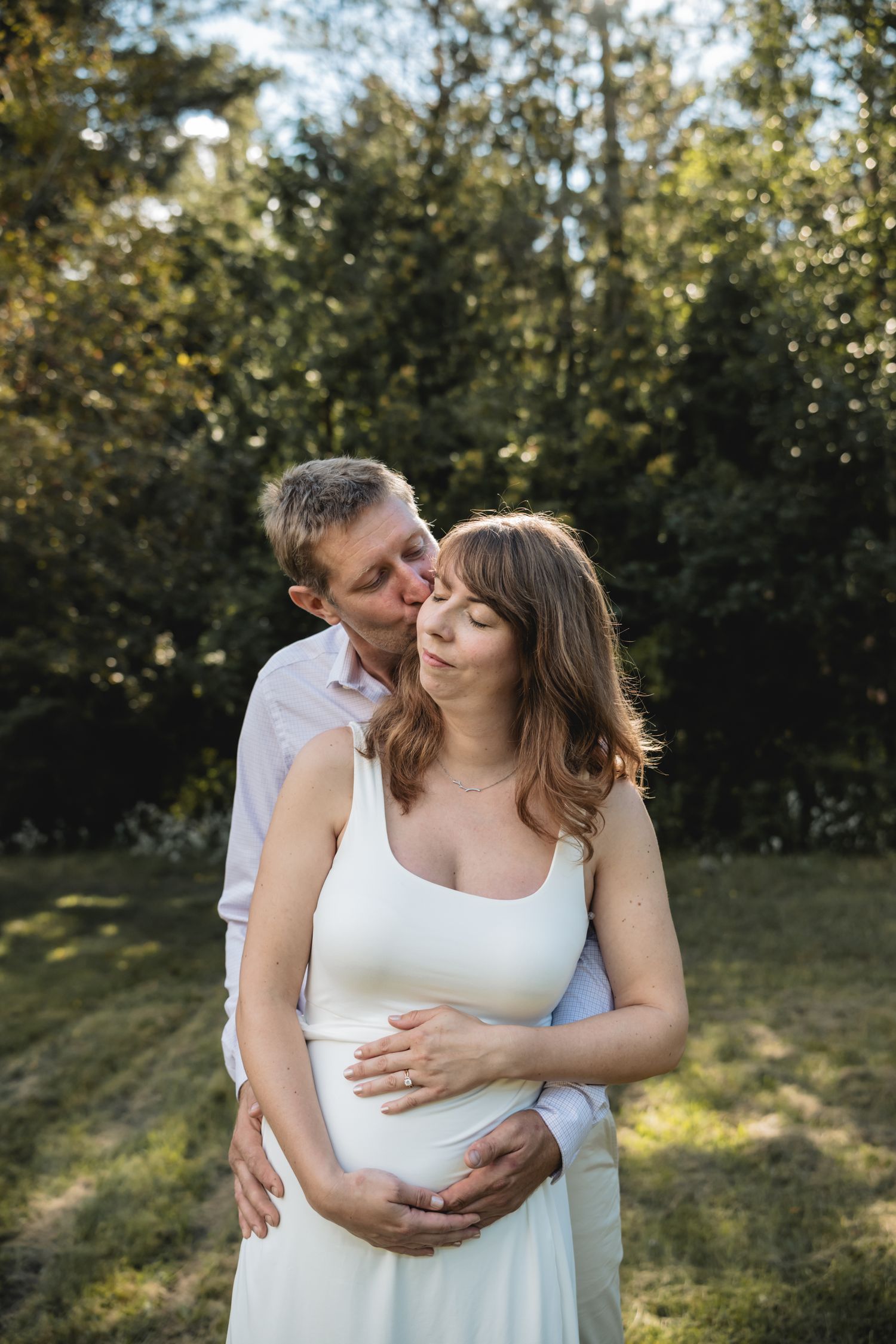 A man kisses a pregnant woman on the cheek while embracing her from behind in a sunlit garden.