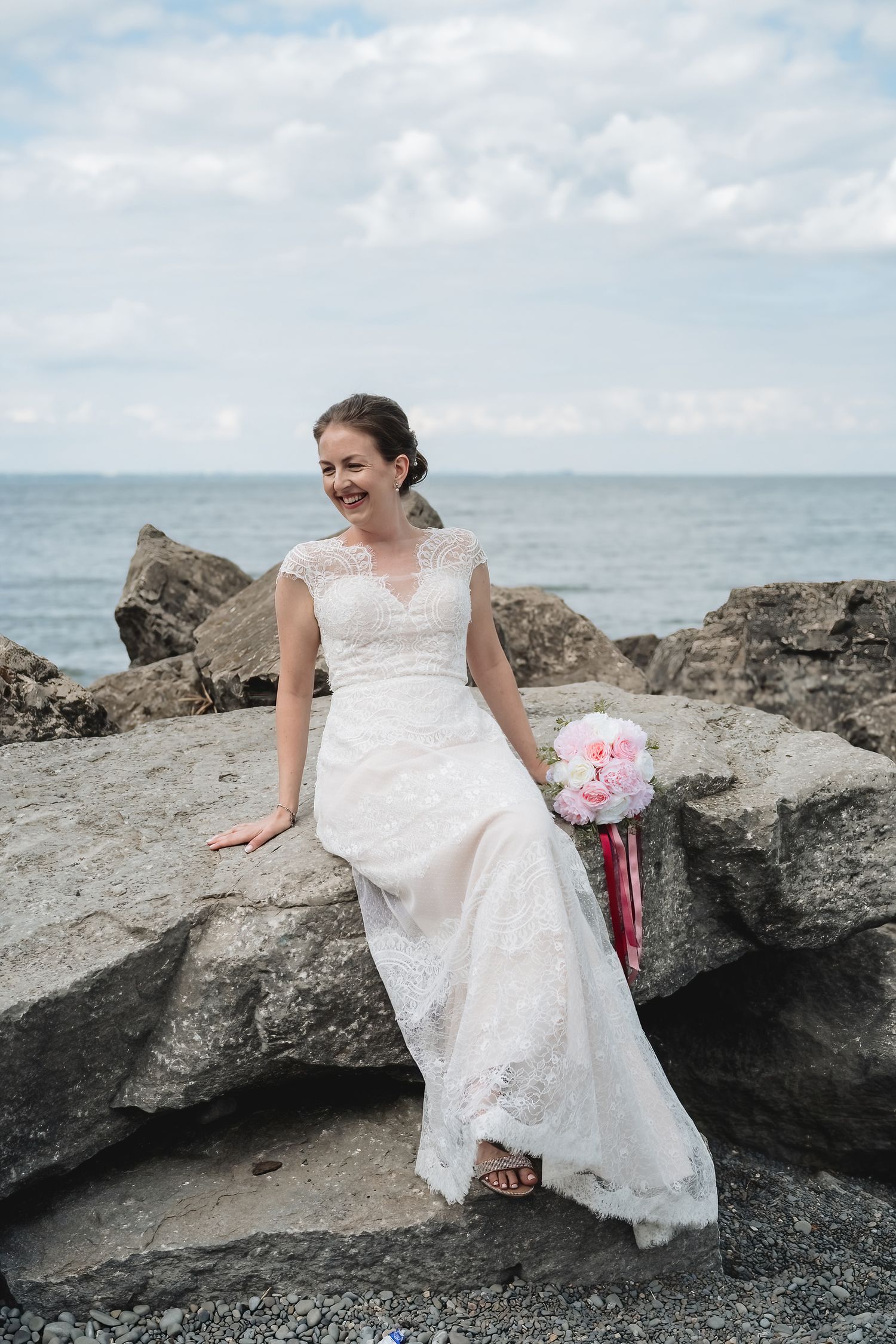 Smiling bride in white lace gown sits on coastal rocks holding pink bouquet with ocean backdrop.