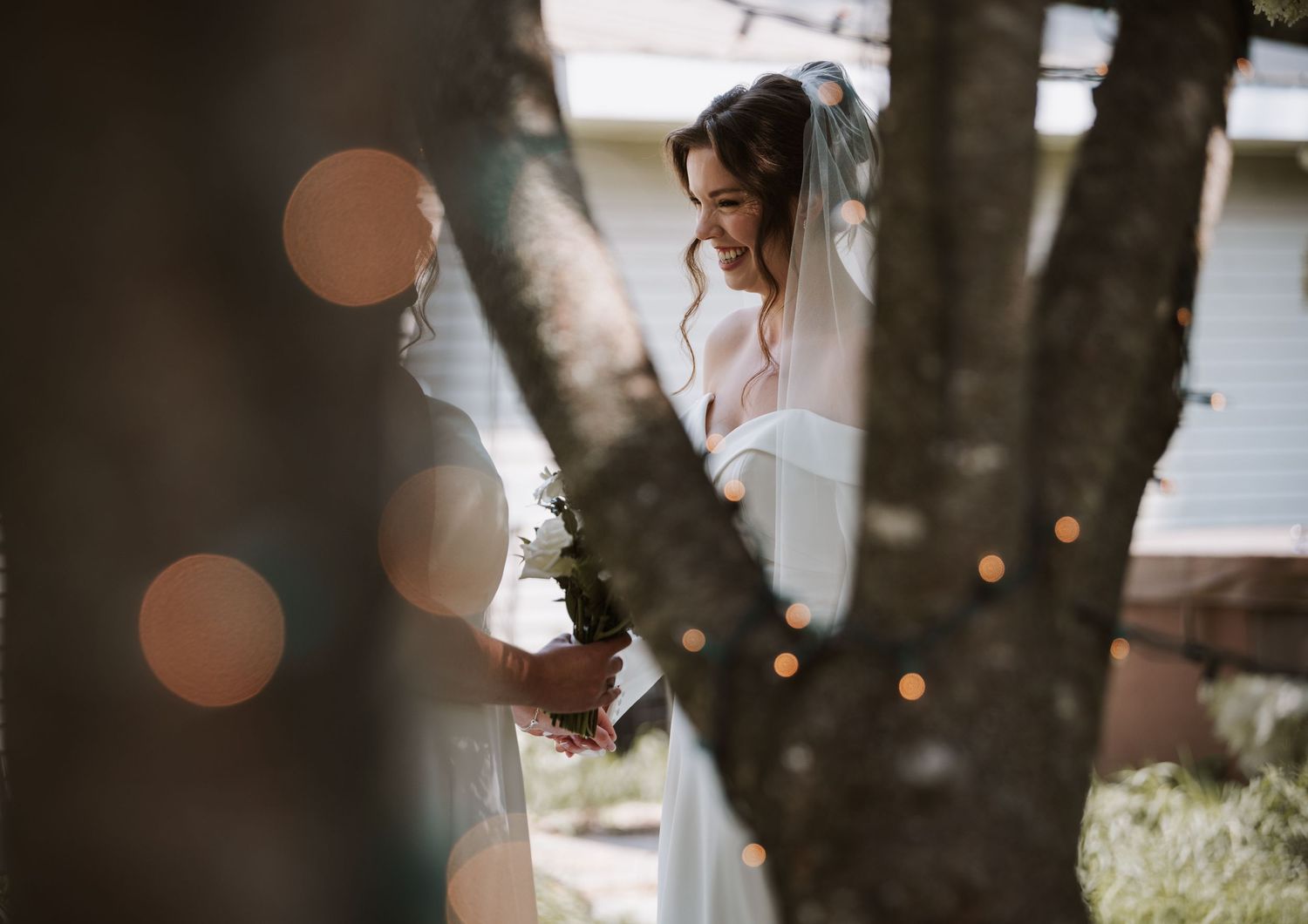 Bride smiling joyfully among trees decorated with fairy lights on her wedding day.