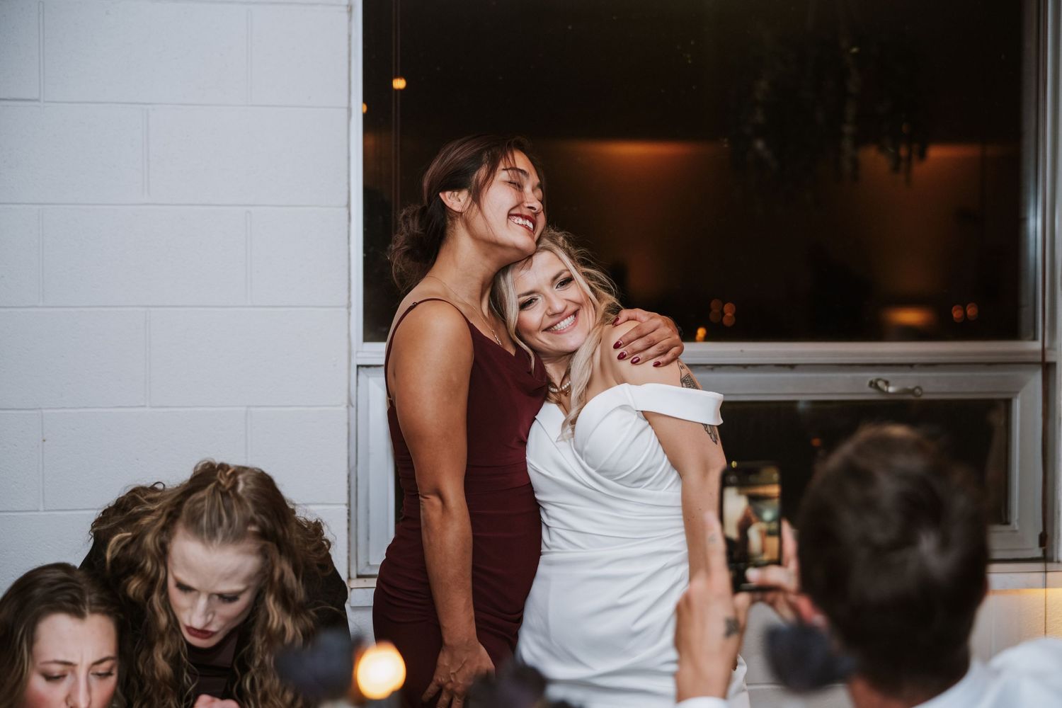 Bride in white dress hugs bridesmaid in burgundy gown while guests celebrate at reception.